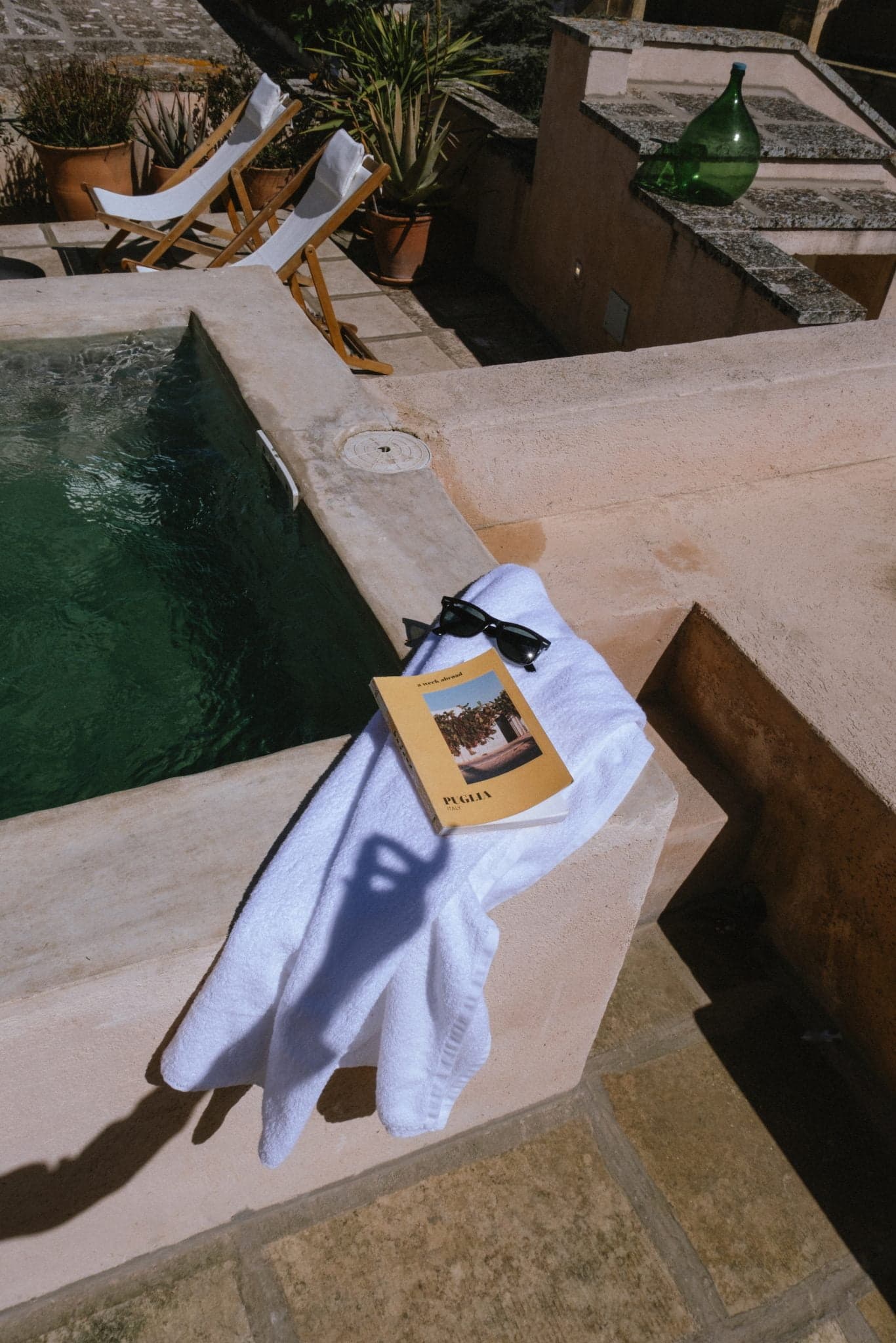Une scène ensoleillée au bord d'une piscine avec une serviette blanche drapée sur le bord, ainsi qu'une paire de lunettes de soleil noires et un livre intitulé "PLAYA". Deux chaises longues vides sont visibles en arrière-plan, ainsi qu'une grande bouteille en verre verte et des plantes en pot. L'eau semble turquoise.