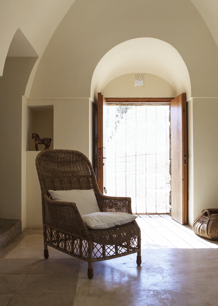 A serene room with vaulted ceilings features a wicker chair with a white cushion positioned near an open wooden door. Sunlight filters through a bamboo curtain hanging at the door. A small animal sculpture is displayed in a niche on the wall, and a woven basket is nearby.
