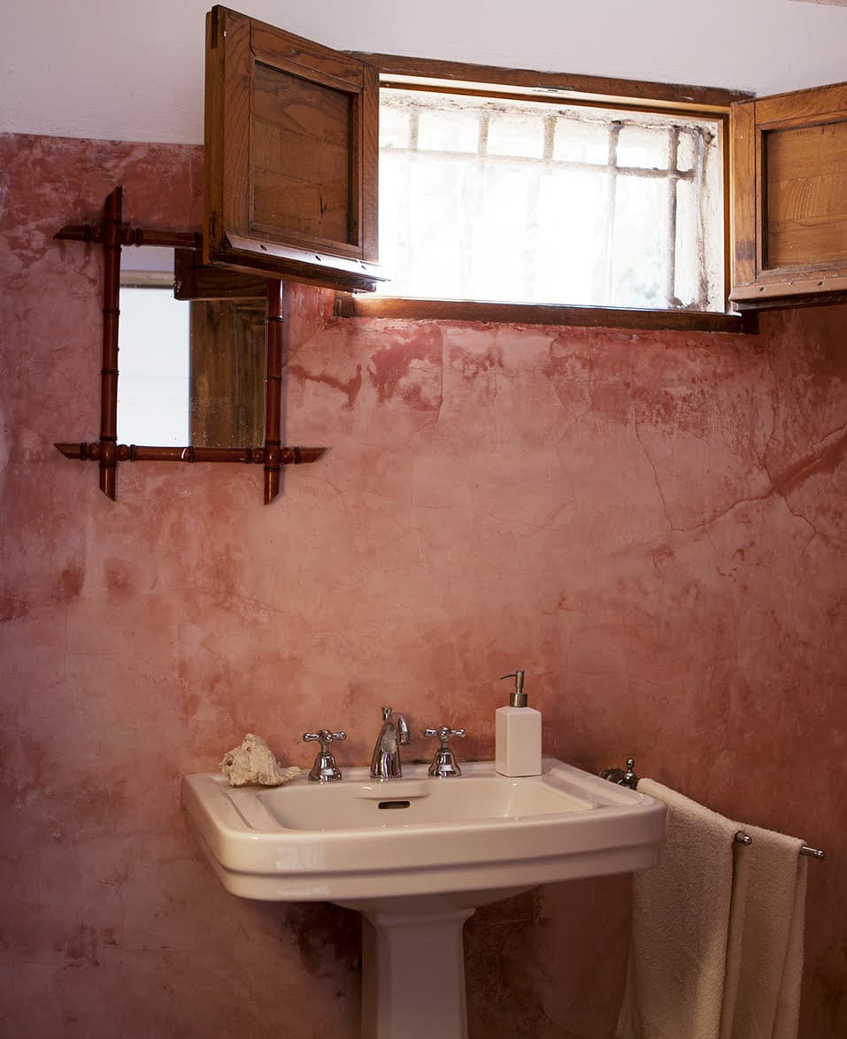 An old-fashioned bathroom features a pedestal sink with a soap dispenser and shell decorations. Above it is a small wooden-framed mirror. A tall window with wooden shutters lets light into the room, which has textured pink-brown walls. A towel is hanging on the right.