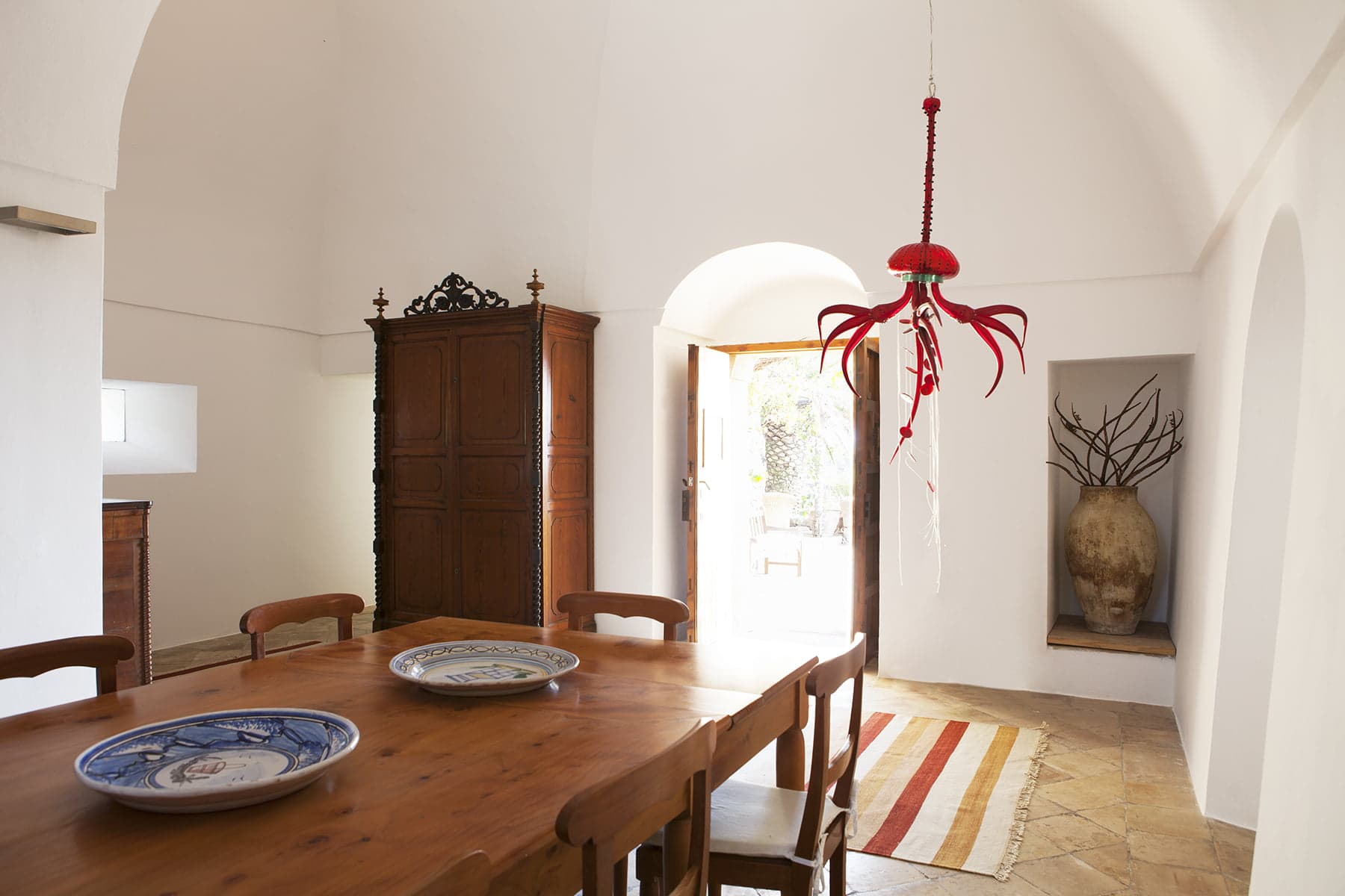 A sunlit dining room features a wooden table with six chairs, adorned with decorative plates. A red chandelier hangs from the ceiling. The room includes an antique wooden cabinet, an arched door, and a beige rug with red stripes. An urn with branches is placed near the door.