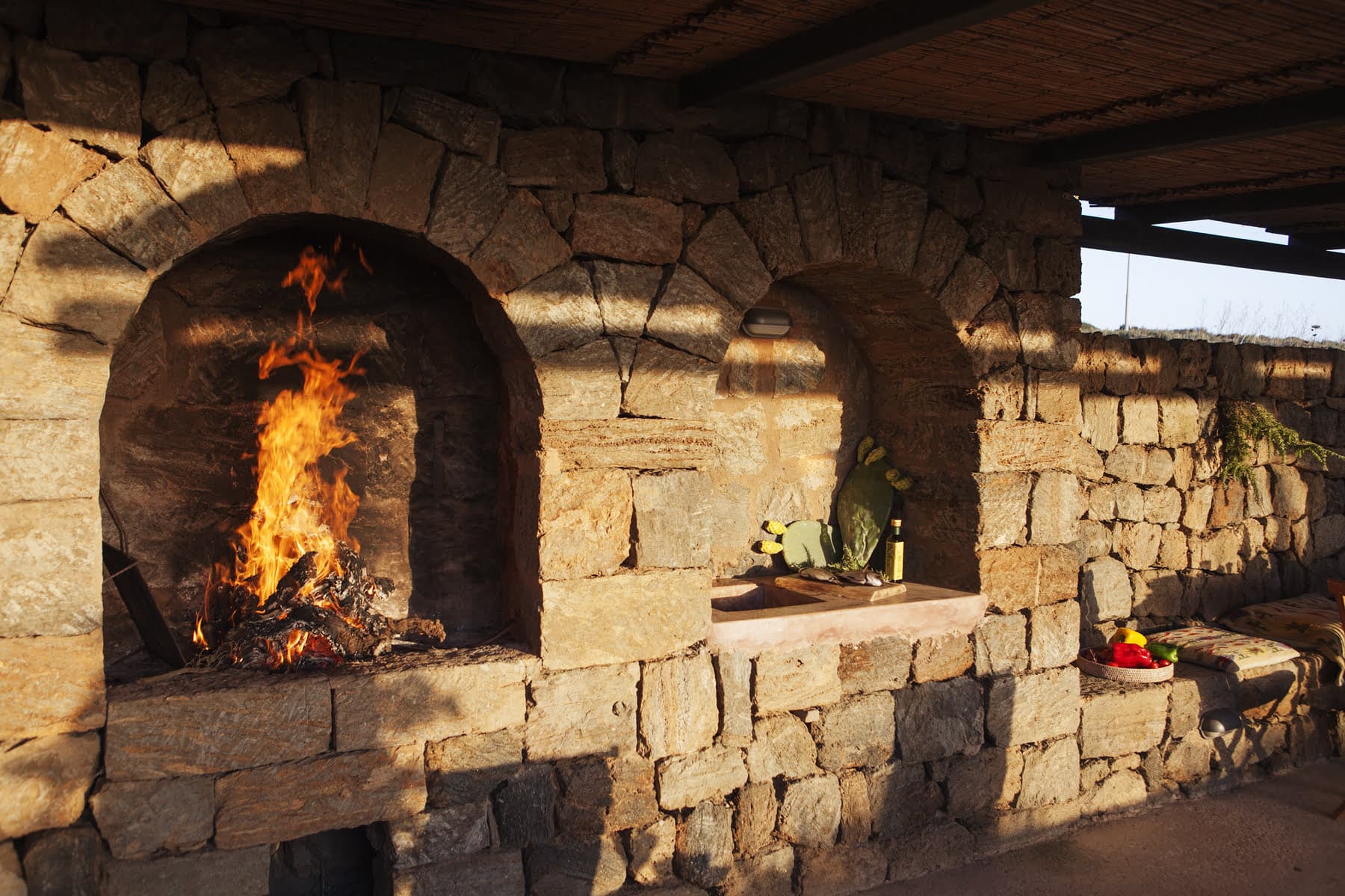 A rustic stone outdoor kitchen with two arched openings. The left opening houses a roaring fire with burning wood. The right side features a stone countertop with various items, including a bottle and some greenery. Sunlight casts shadows on the stone walls.