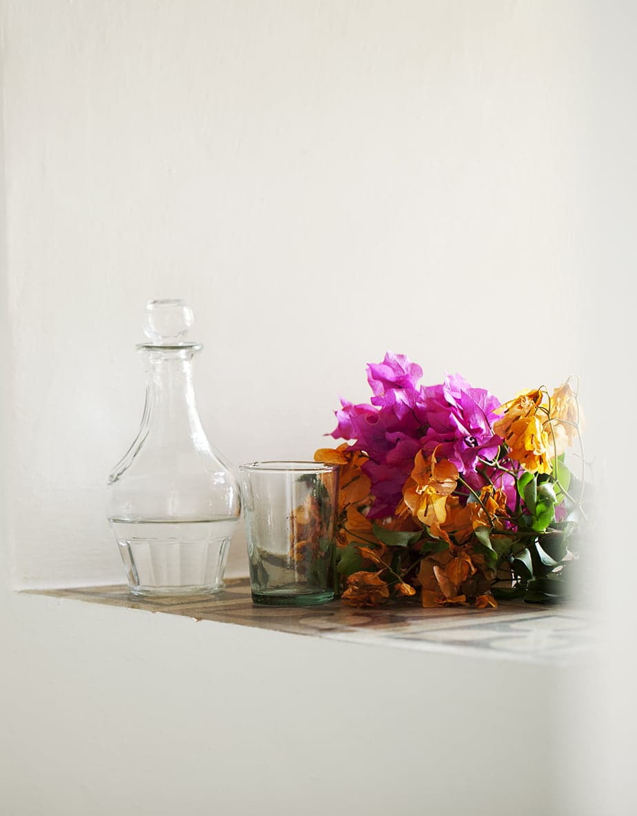 A clear glass pitcher and an empty glass are placed on a marble shelf next to a vibrant bouquet of pink, purple, and orange flowers against a solid white wall.
