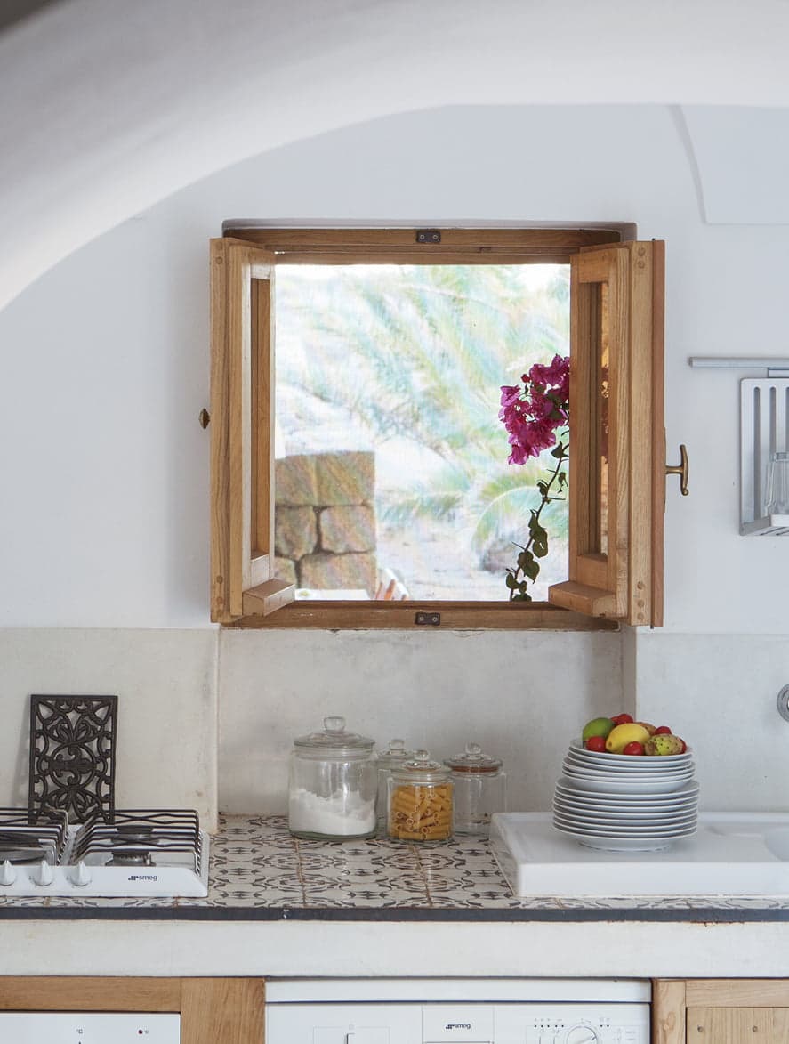 A cozy kitchen with a small wooden-framed window, partially open, revealing a view of greenery and flowers outside. Inside, there is a countertop with jars, plates, and a bowl of fruit. The backsplash features patterned tiles, and a gas stove is visible on the left.