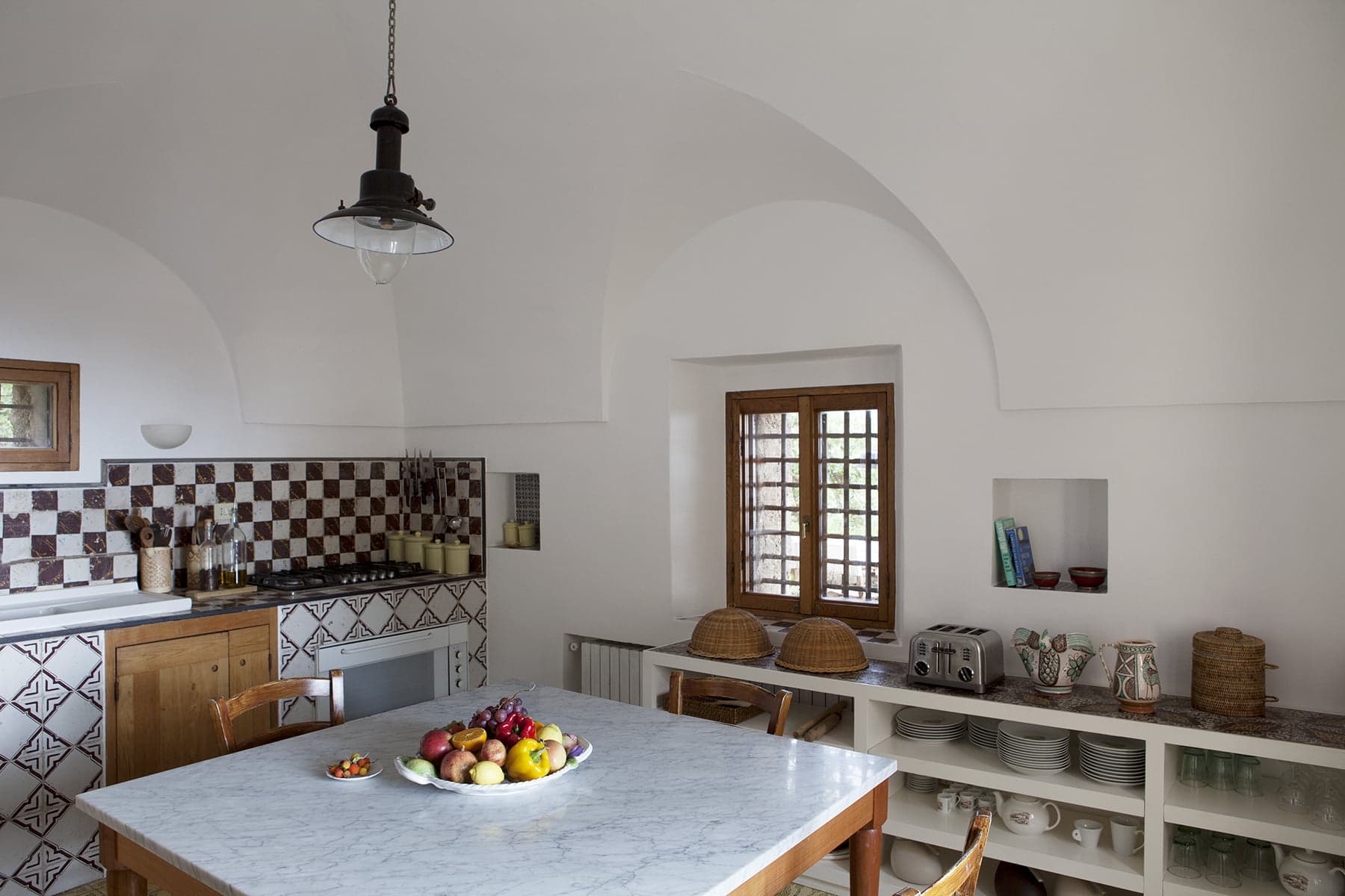 A bright kitchen with white walls and a vaulted ceiling features a marble table with a bowl of fruit, wooden cabinets, a tiled backsplash, open shelves with dishware, a toaster, wall shelves filled with books, and a hanging pendant light.