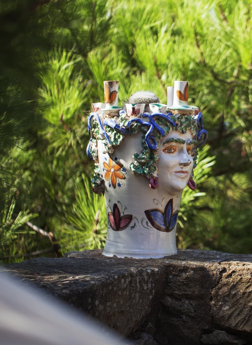 A ceramic vase shaped like a human head, adorned with floral patterns and intricate vines, sits on an outdoor stone ledge. The background features lush green foliage, suggesting a garden or natural setting.