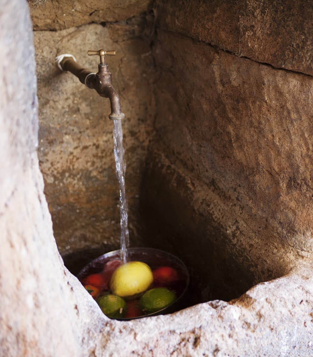A stone sink with water flowing from a bronze faucet. In the sink, a metal bowl holds various fruits, including a lemon and a few tomatoes, being washed by the running water. The sink is surrounded by rustic stone walls.