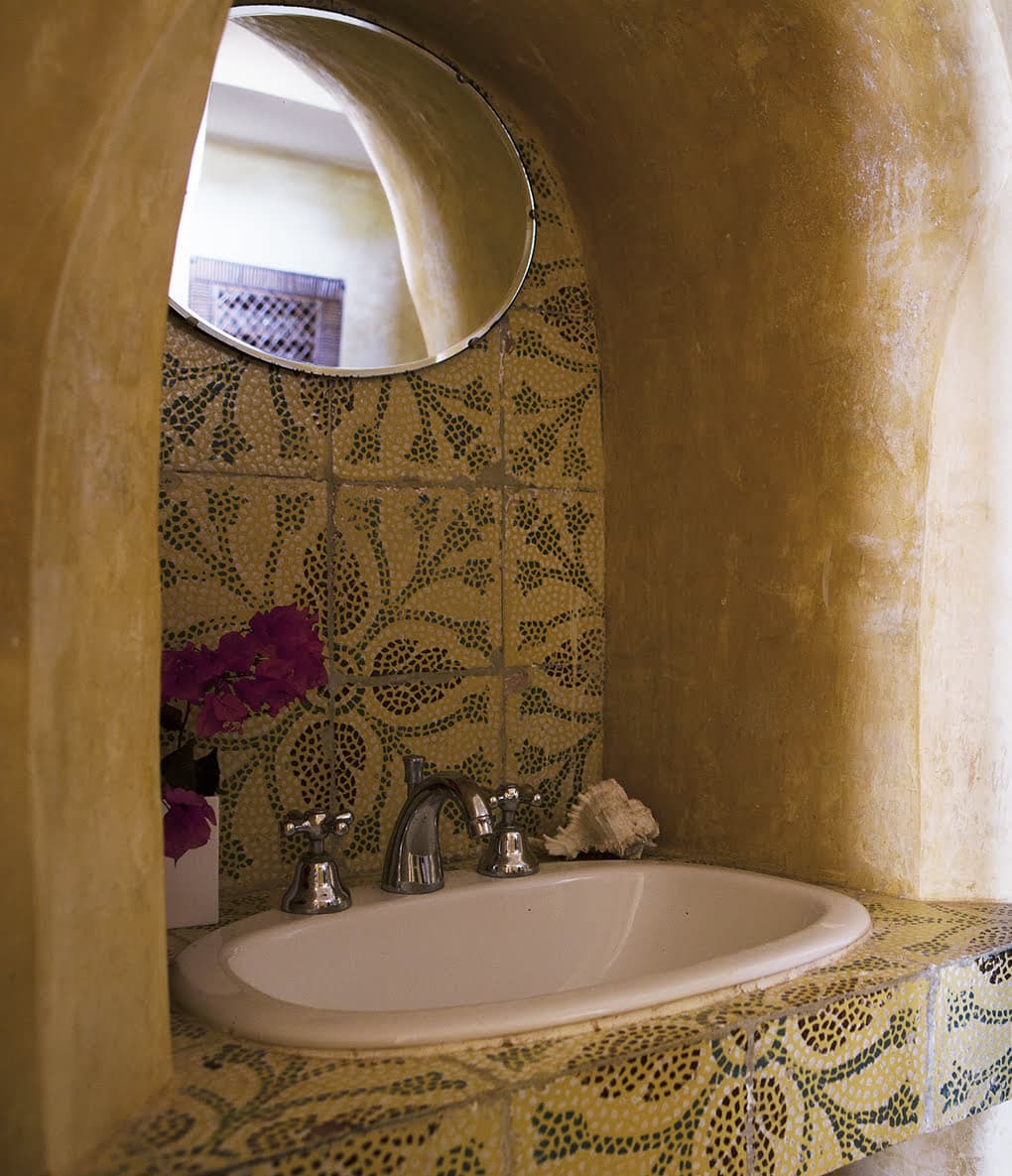 A rustic bathroom featuring a circular mirror above a ceramic sink. The countertop and backsplash of the sink are adorned with intricate green and yellow mosaics. A vase with vibrant purple flowers and a seashell are placed beside the sink. The arched alcove adds depth to the space.