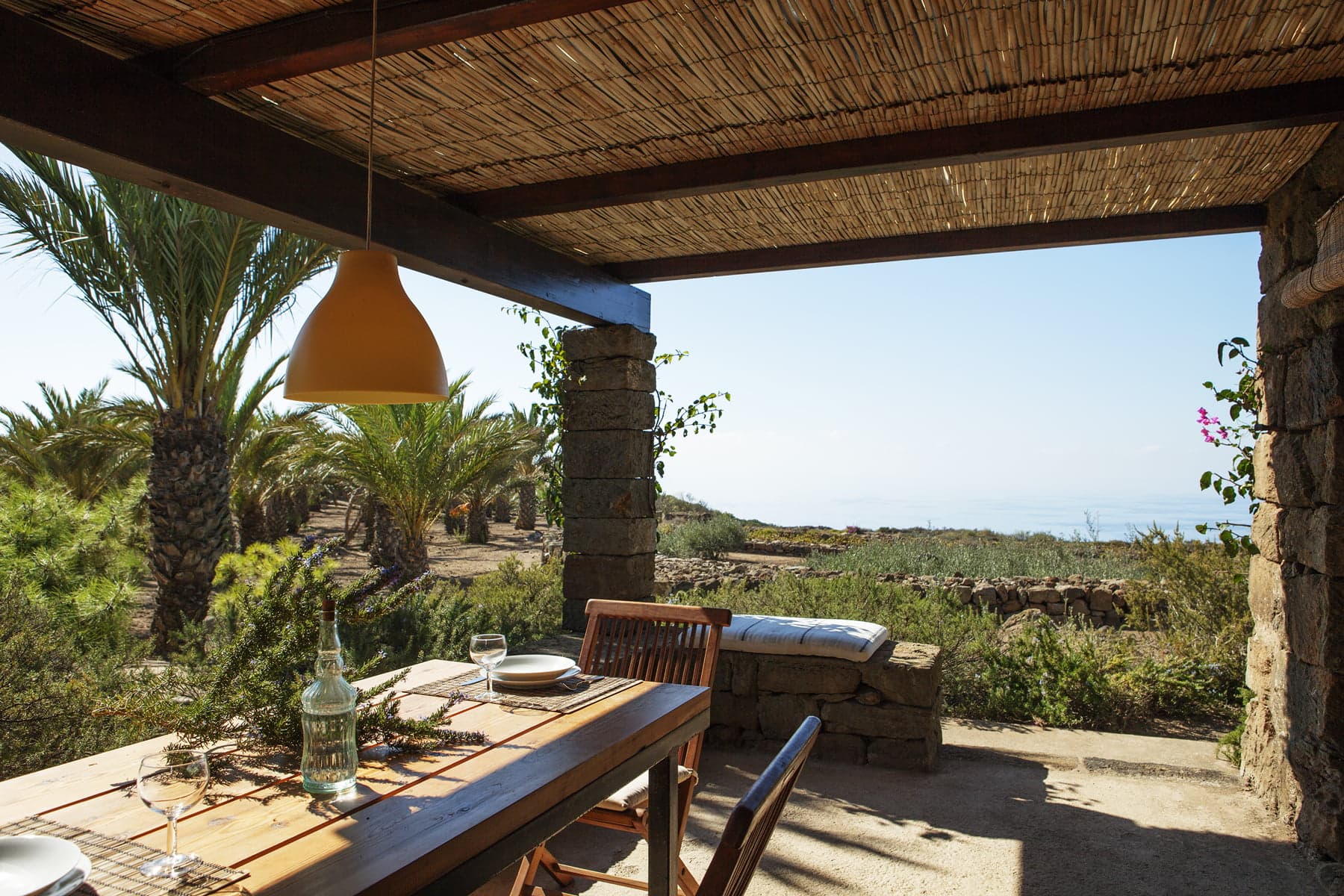 A covered outdoor dining area with a wooden table set for a meal, wooden chairs, and a yellow pendant light. The view includes a lush green garden with palm trees and a distant view of the sea under a clear blue sky.