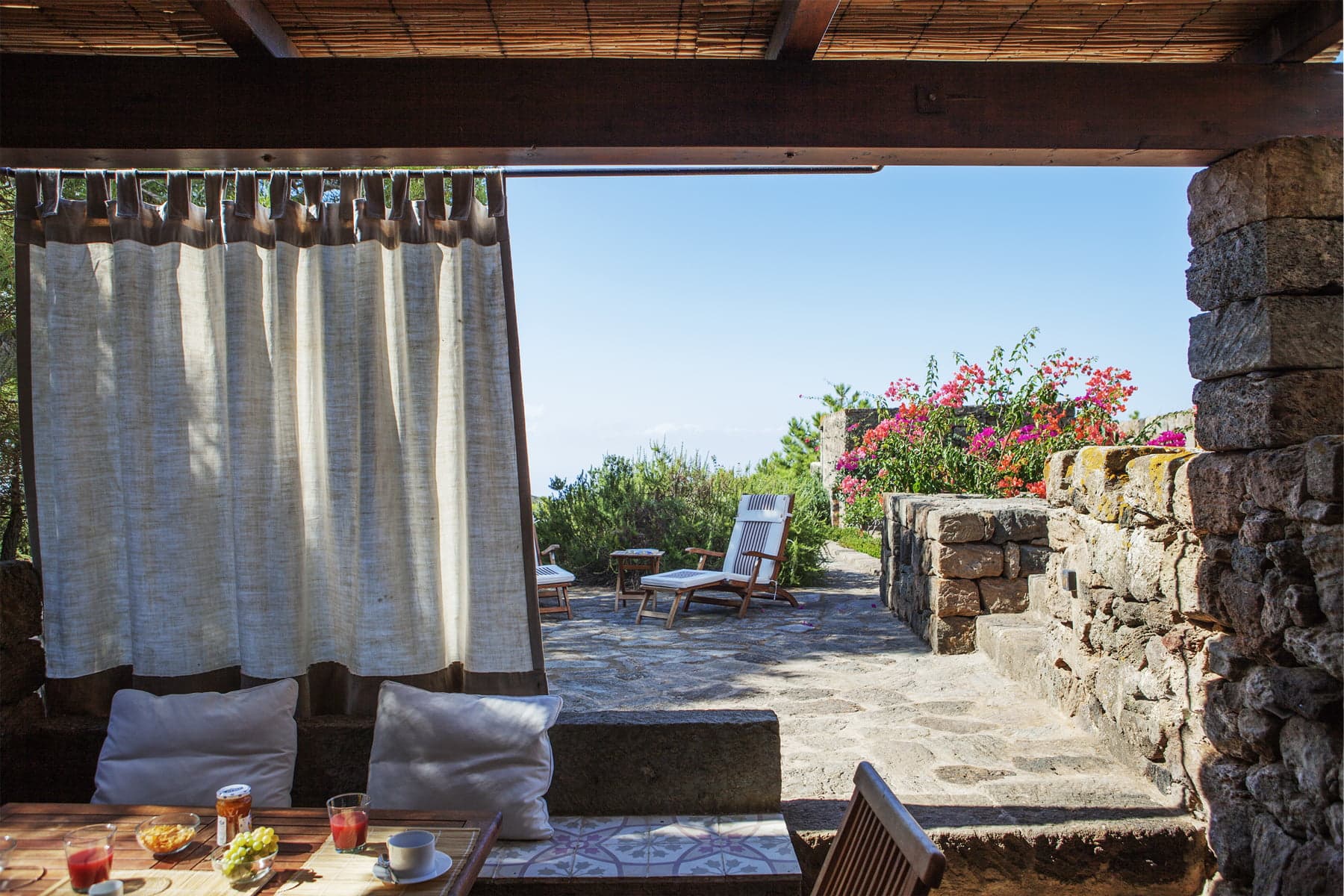 A cozy outdoor patio features a rustic stone wall and a tiled floor. It is furnished with wooden chairs and tables set for breakfast with drinks and food. Beyond the patio, there are additional wooden lounge chairs, brightly colored flowers, and a panoramic view of greenery and the sky.