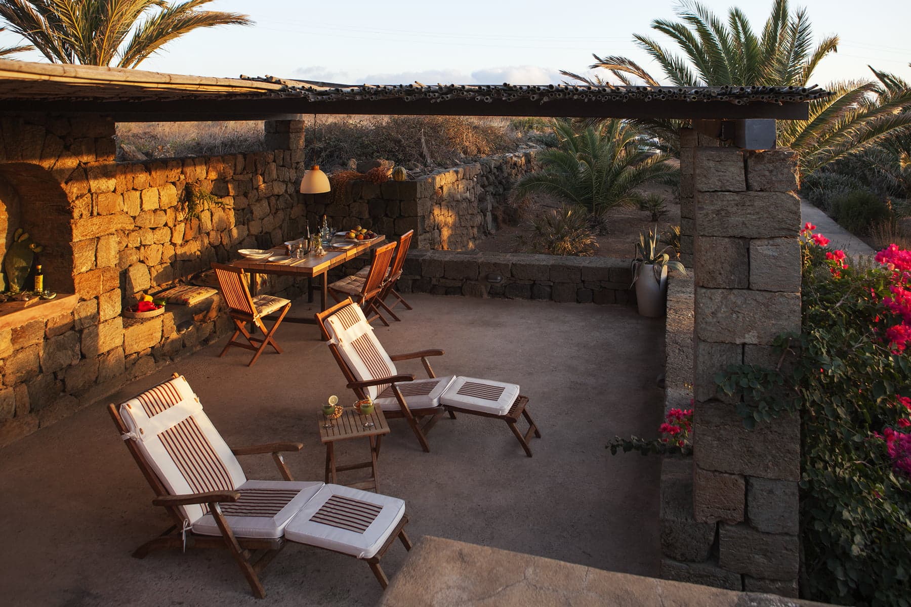 A serene outdoor patio with two wooden loungers topped with white cushions, a small wooden side table, a dining area with a table and chairs, all backed by stone walls. Tropical plants and blooming flowers frame the scene, with palm trees in the background.