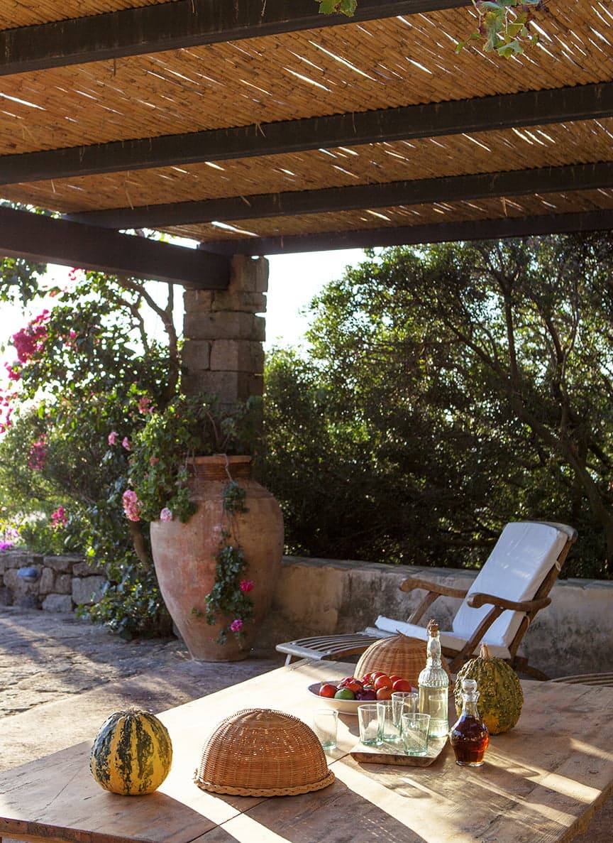 A rustic outdoor terrace features a wooden pergola covered with straw mats, a large clay pot with flowering plants, and a wooden table adorned with melons, glassware, and a covered basket. A white lounge chair is set against a backdrop of lush greenery.