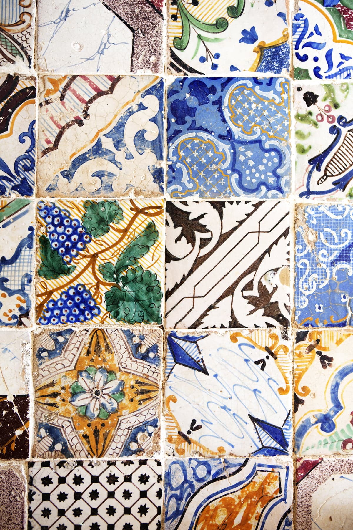 A mosaic of colorful ceramic tiles with intricate patterns, each featuring unique designs and illustrations. The tiles showcase a variety of colors, including blue, green, yellow, and white, with a blend of floral, geometric, and abstract motifs.
