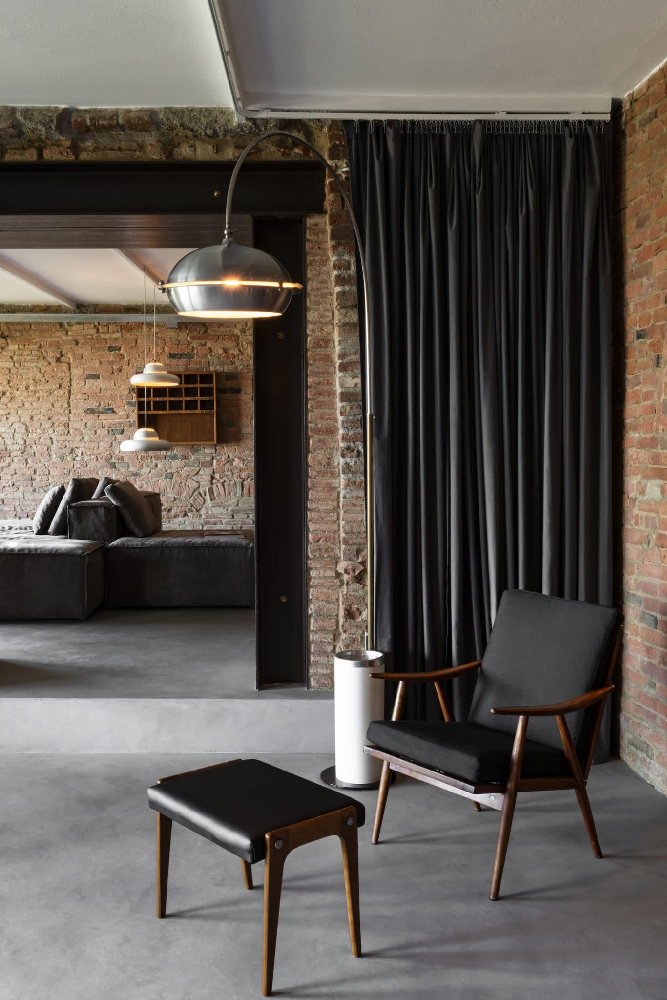 A modern living space with exposed brick walls and a gray floor. It features a black armchair with a matching ottoman, a small side table, and an industrial-style pendant light. A dark curtain partially conceals a door leading to another room with additional seating.