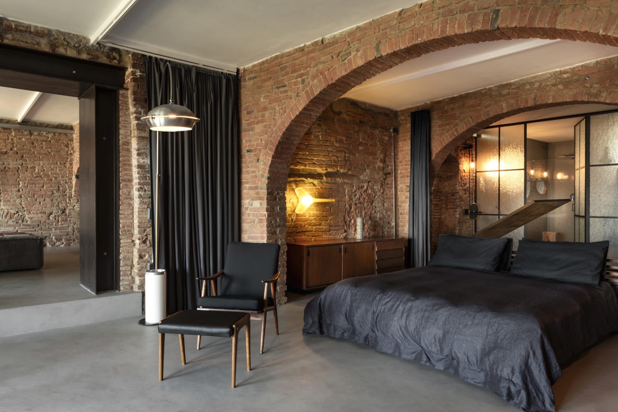 Une chambre moderne aux murs en briques apparentes comprend un lit double avec une literie sombre, une chaise noire aux accents dorés, une commode en bois et un grand lampadaire. Les arcades et l'éclairage d'ambiance ajoutent de la profondeur, tandis qu'un rideau partiellement ouvert mène à un espace adjacent.