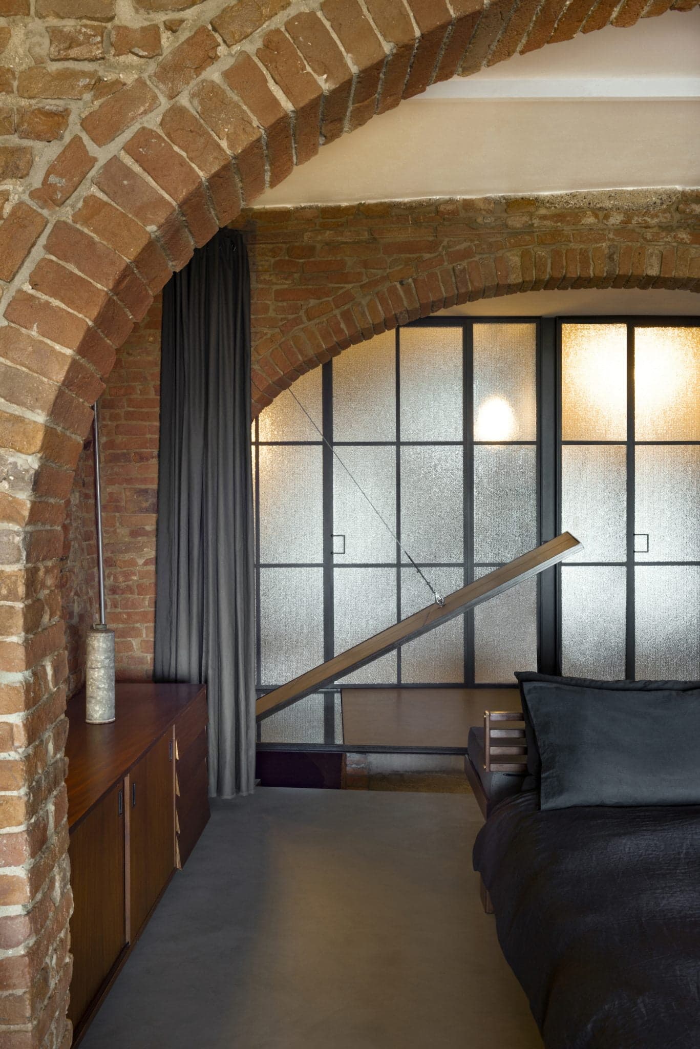 A bedroom with rustic exposed brick arches, a large frosted glass window, and modern decor. The bed features dark bedding, and there is a wooden side table with a vase. A gray curtain is hung beside the window, and an elegant wooden wardrobe is situated beneath the arch.