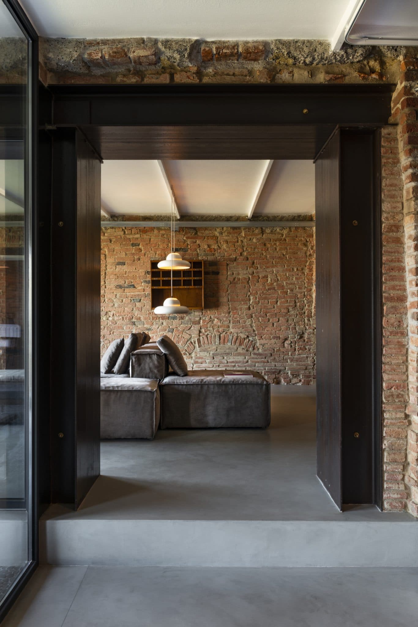 A modern interior with a minimalist design, featuring exposed brick walls and a concrete floor. A gray sectional sofa is centered beneath two white pendant lights. A wooden bookshelf is mounted on the brick wall behind the sofa.