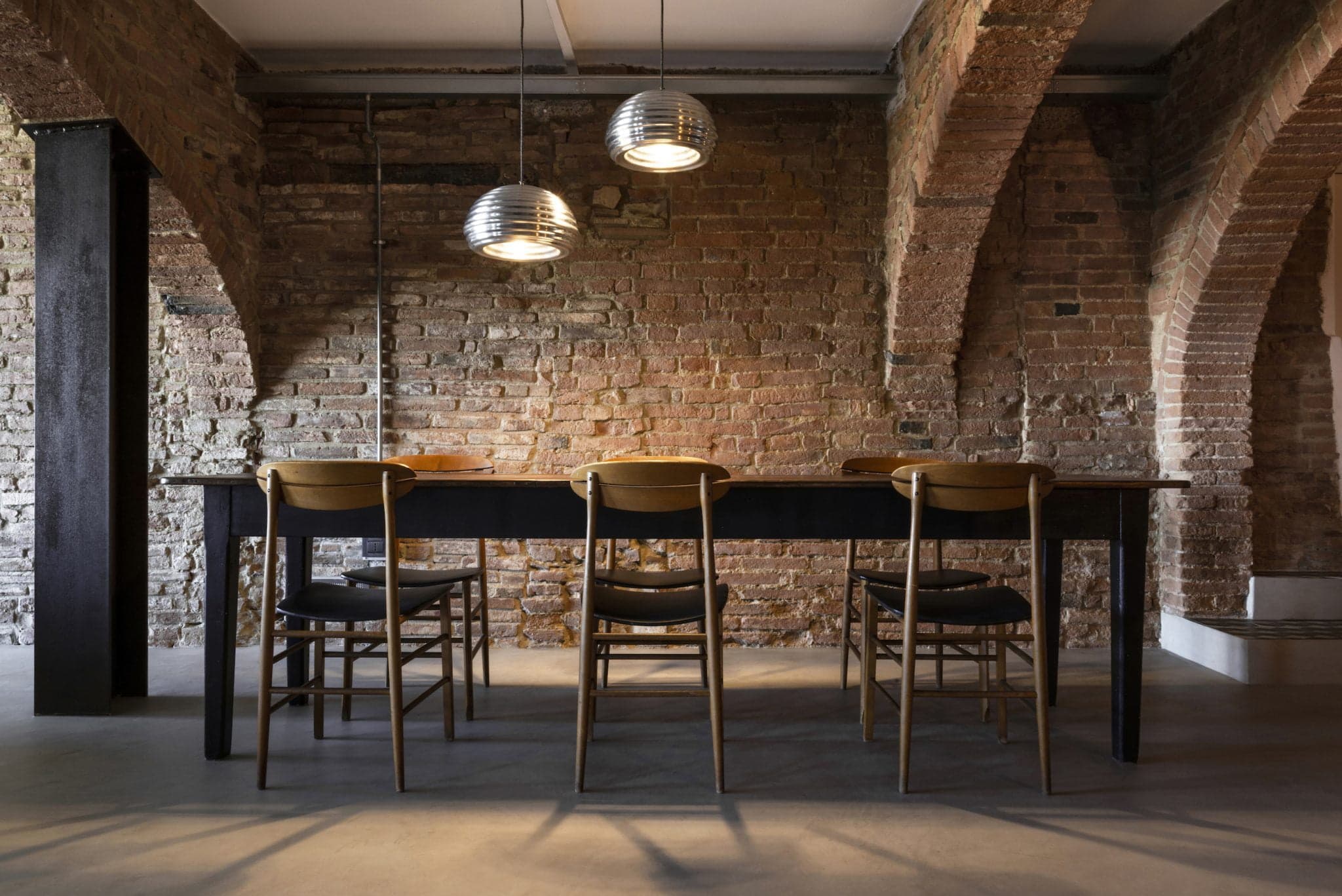 Un coin repas avec une table en bois rustique et cinq chaises assorties. La pièce est dotée de murs en briques apparentes et de plafonds voûtés, ce qui lui donne un aspect industriel. Deux suspensions modernes sont suspendues au-dessus de la table, projetant une lueur chaleureuse sur la scène.
