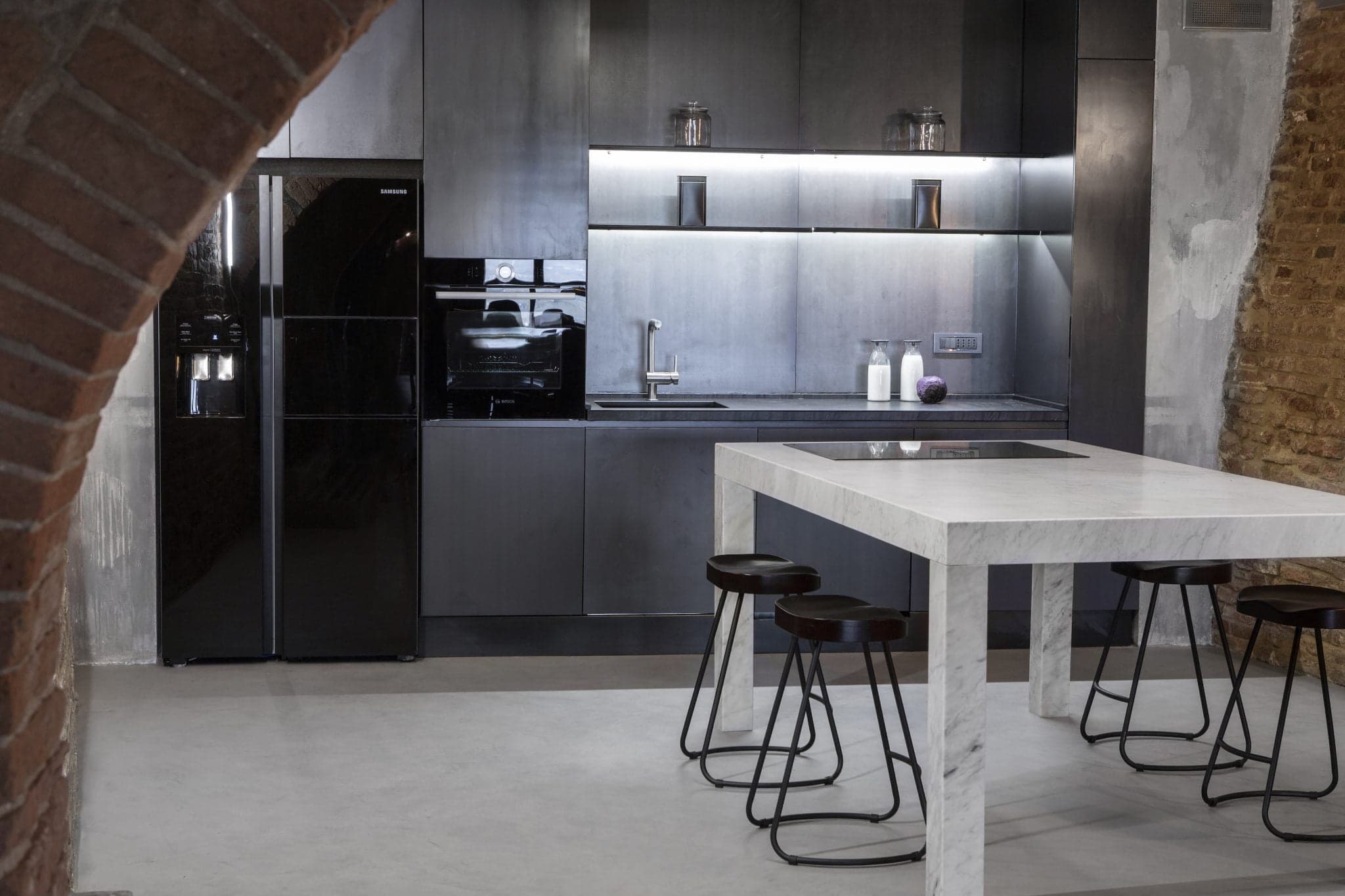 Une cuisine moderne avec des meubles sombres et un réfrigérateur noir. Un élégant îlot en marbre blanc avec quatre tabourets de bar noirs se trouve au premier plan. L'espace présente un mélange d'éléments en métal et en bois, avec une arche en briques apparentes sur le côté gauche de l'image.
