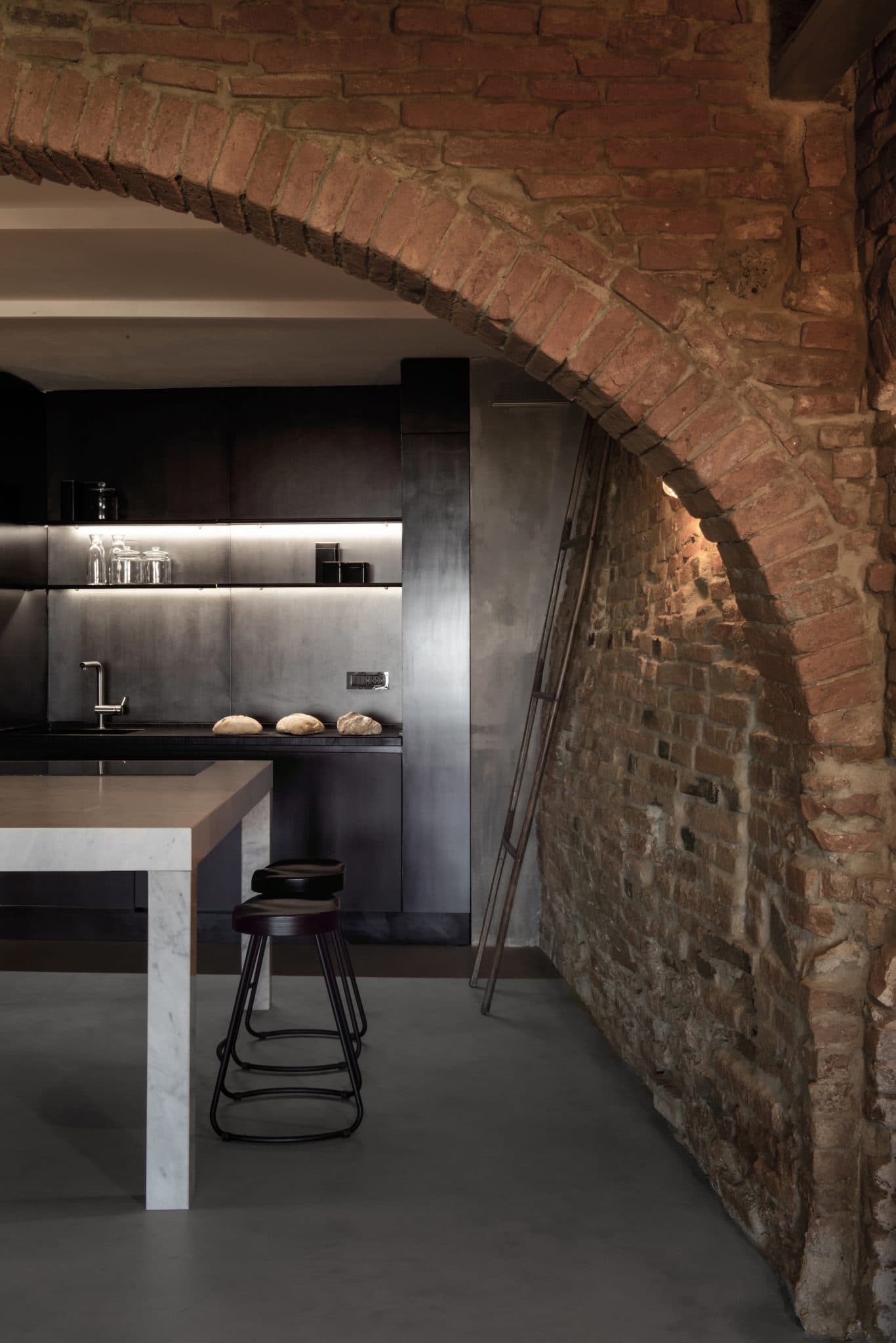 A modern kitchen featuring a rustic, vaulted brick wall. The space includes dark cabinets, a countertop, minimalist decor, and a stainless steel sink. A large metal ladder leans against the brick wall. In the foreground, there is a white island countertop with a black stool.