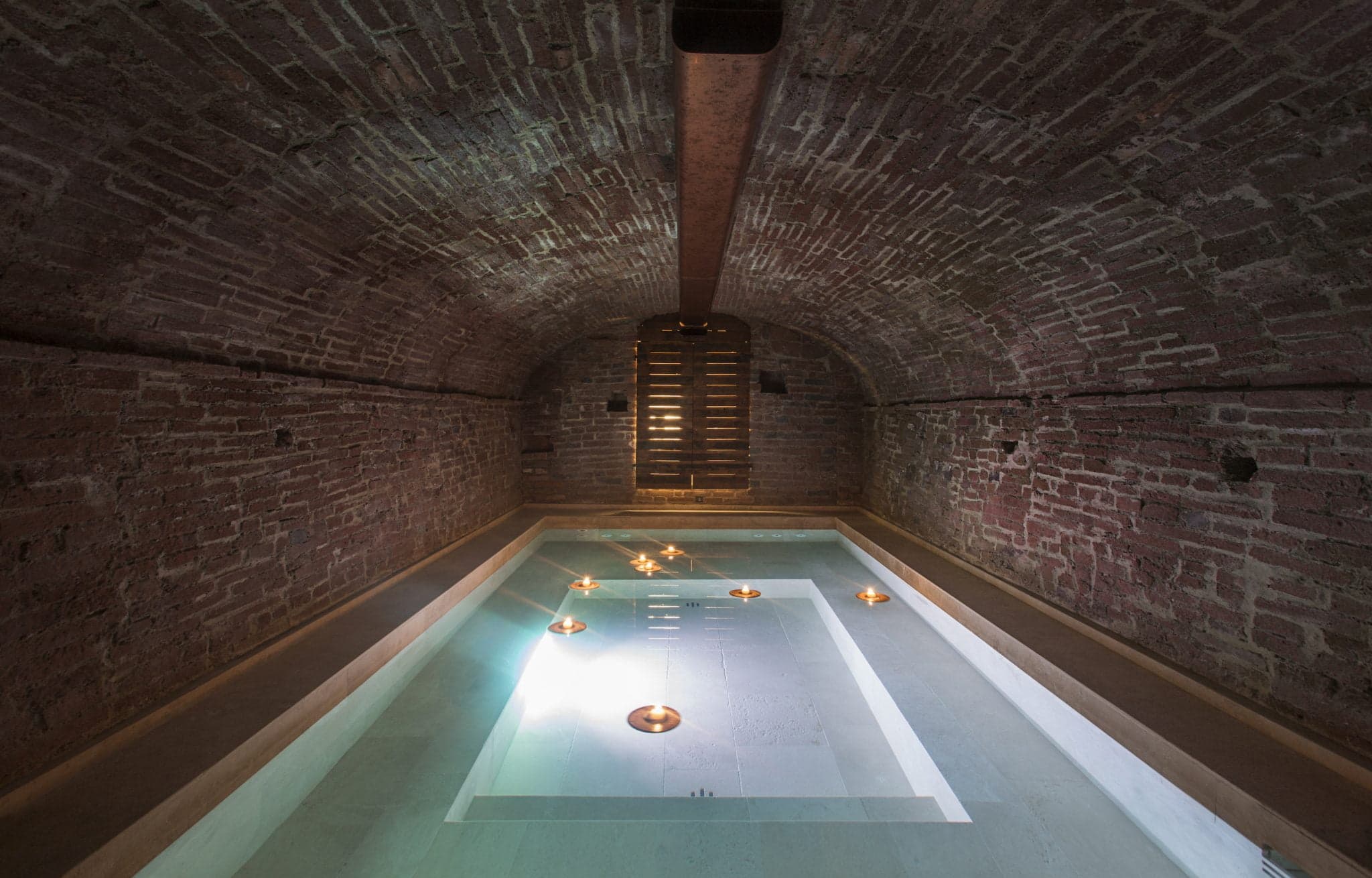The image shows an underground pool bathed in dim light, nestled within a vaulted brick ceiling structure. Several small floating candles cast a warm, soothing glow across the water, illuminating the space with a tranquil ambiance.
