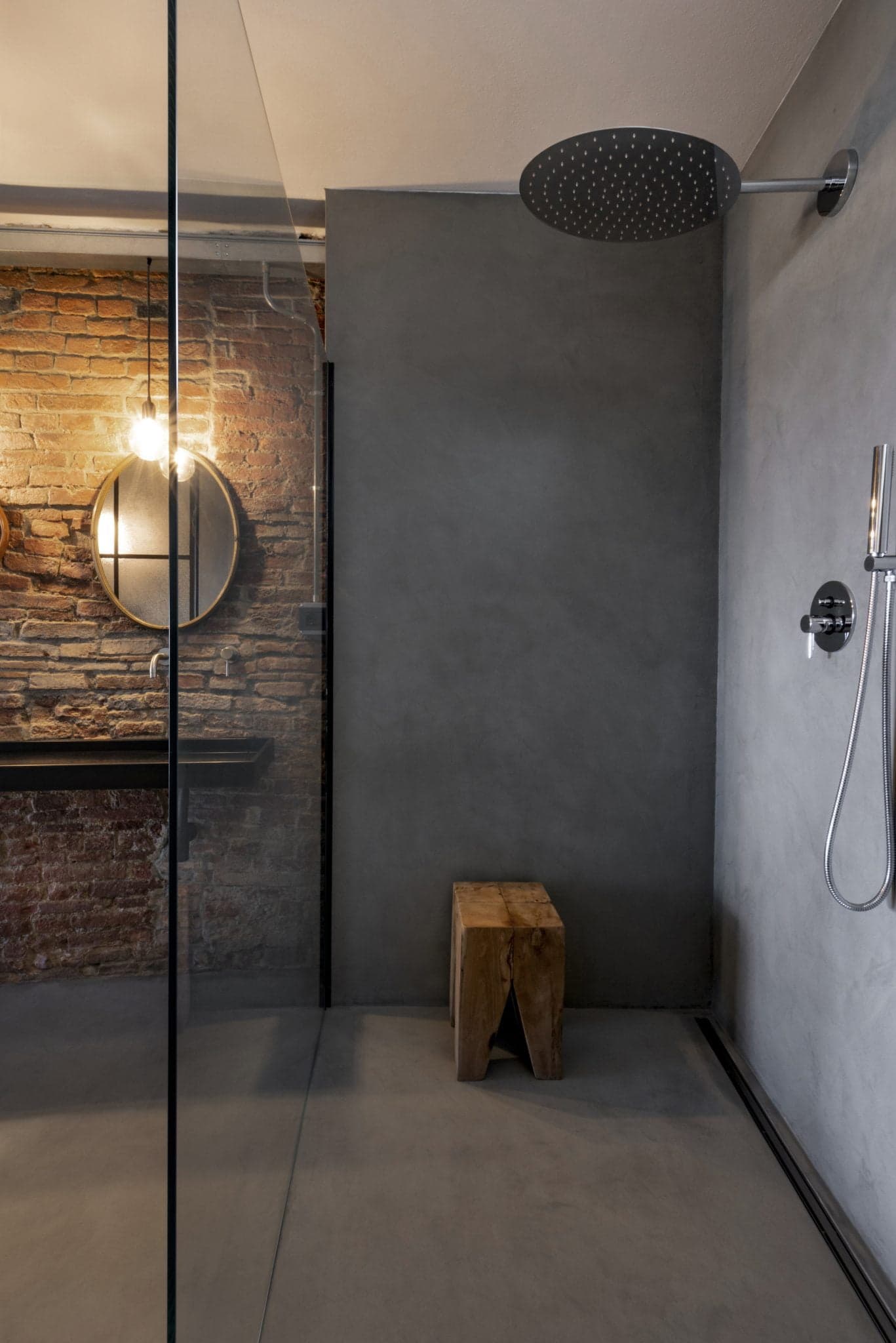 A modern bathroom with a glass-enclosed shower. The shower features a large round rain showerhead mounted on the ceiling. There's a small wooden stool in the corner. To the side, there is a vanity with an exposed brick accent wall, a round mirror, and two lights.