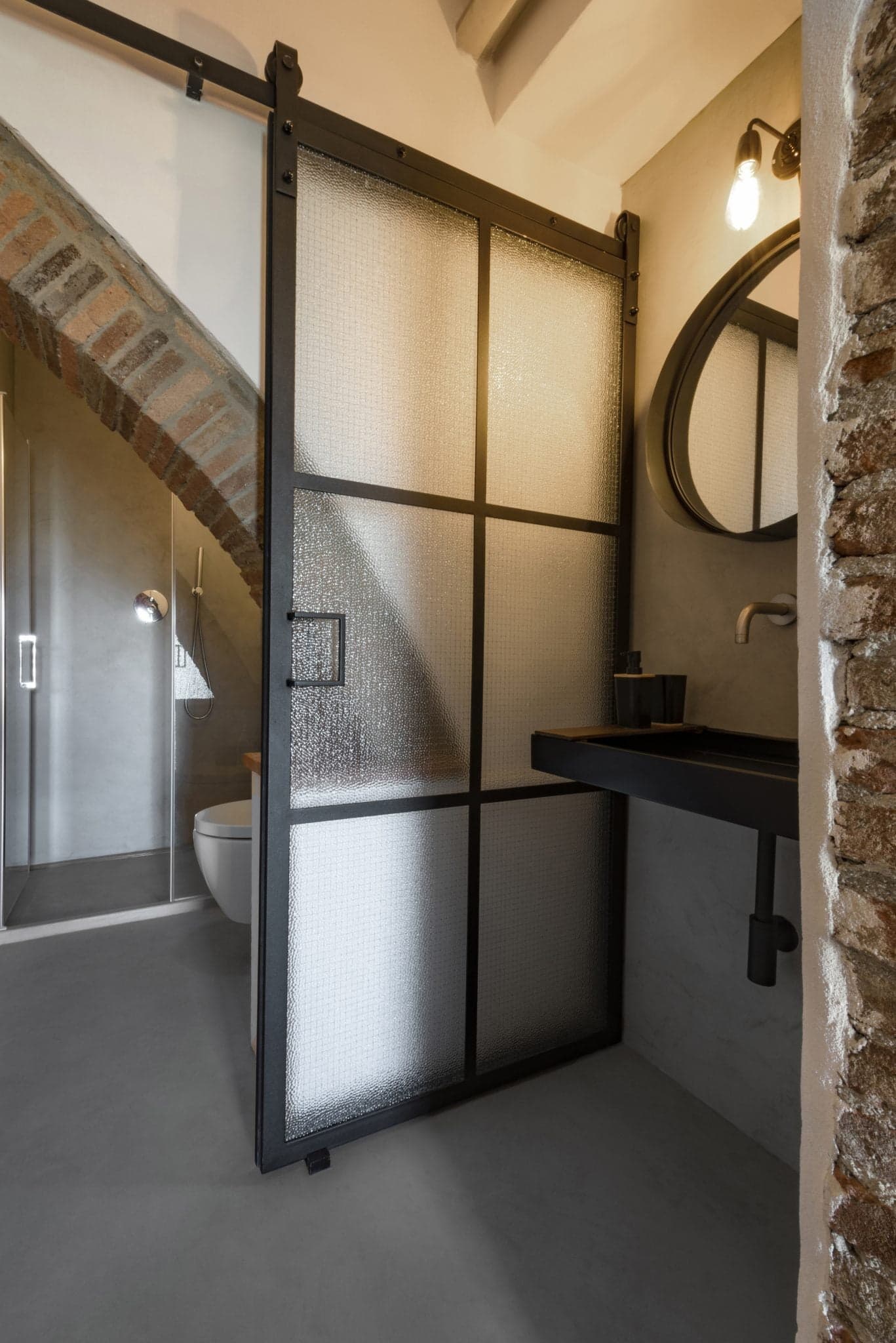 A modern bathroom with a partially open frosted glass sliding door. Inside, the space features rustic brick arches, a dark metal frame around the glass door, a round mirror above a black vanity with a brushed metal faucet, and exposed plumbing.