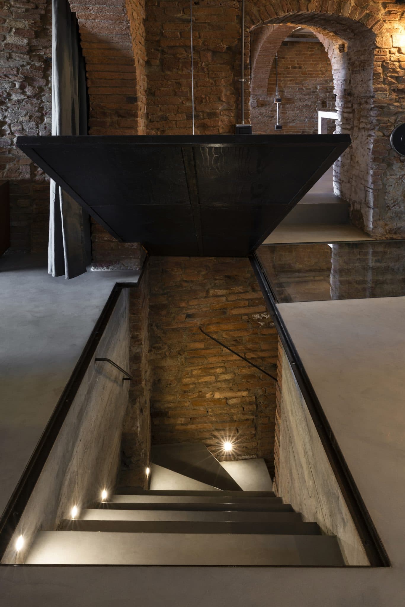 Un escalier étroit et moderne éclairé par de petites lumières descend vers un niveau inférieur avec des murs en briques apparentes, des sols en béton et des arcades courbes, créant une esthétique à la fois industrielle et historique. L'espace est peu meublé et présente un mélange d'éléments architecturaux anciens et nouveaux.