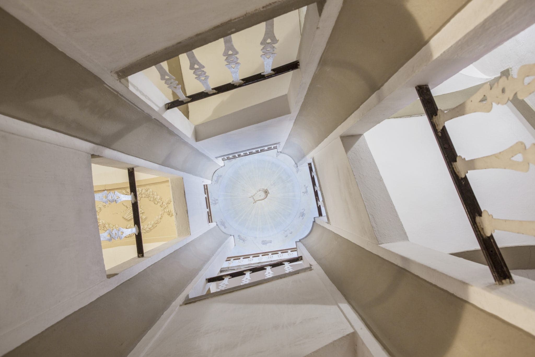 View of a multi-story spiral staircase with wide railings and ornate support beams, leading up to a decorative circular ceiling with intricate patterns. The perspective creates a geometric pattern, with soft lighting highlighting the architectural details.