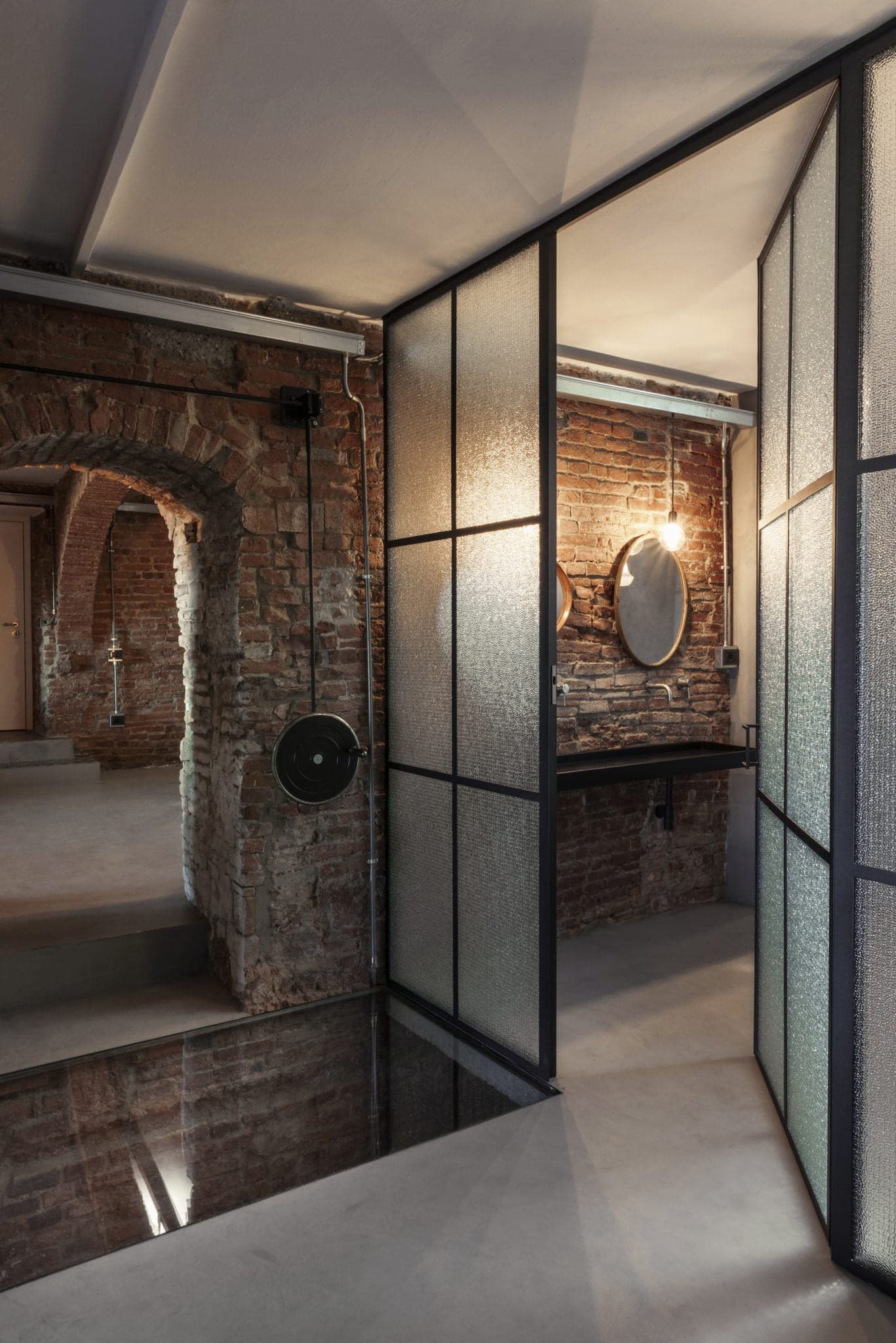 A modern bathroom with rustic brick walls and industrial-style glass partitions with metal frames. The vanity features a round mirror and a wall-mounted faucet. Soft, warm lighting creates a cozy atmosphere, contrasting with the rough texture of the masonry.