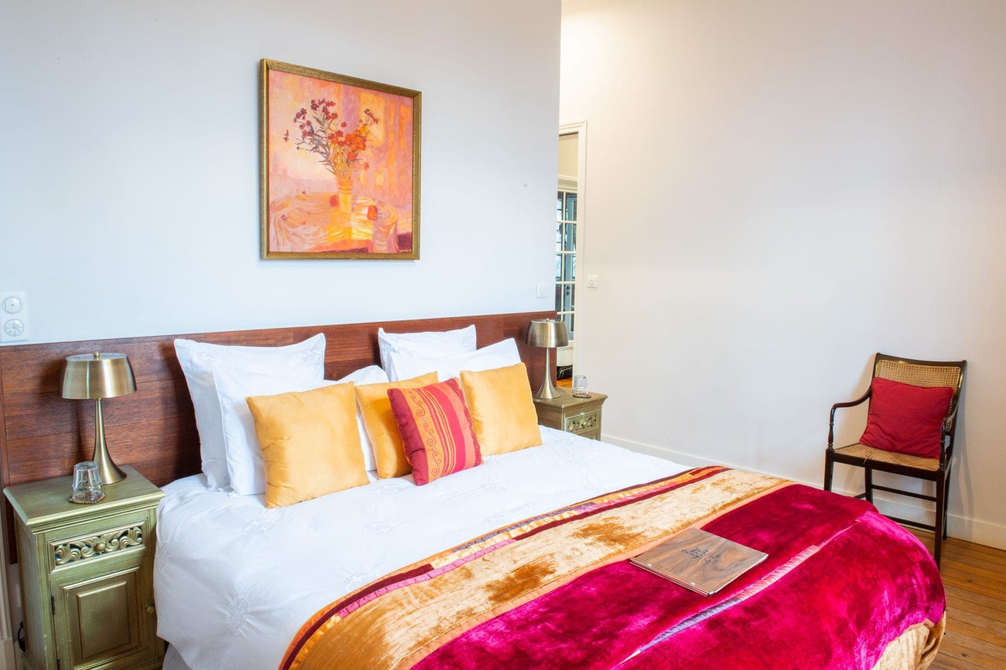 A comfortable room with a double bed and white and orange pillows. The bed is covered with a red and gold blanket. Two vintage-style bedside tables with lamps frame the bed. A colorful painting hangs above the bed. There's a wooden chair in the corner.