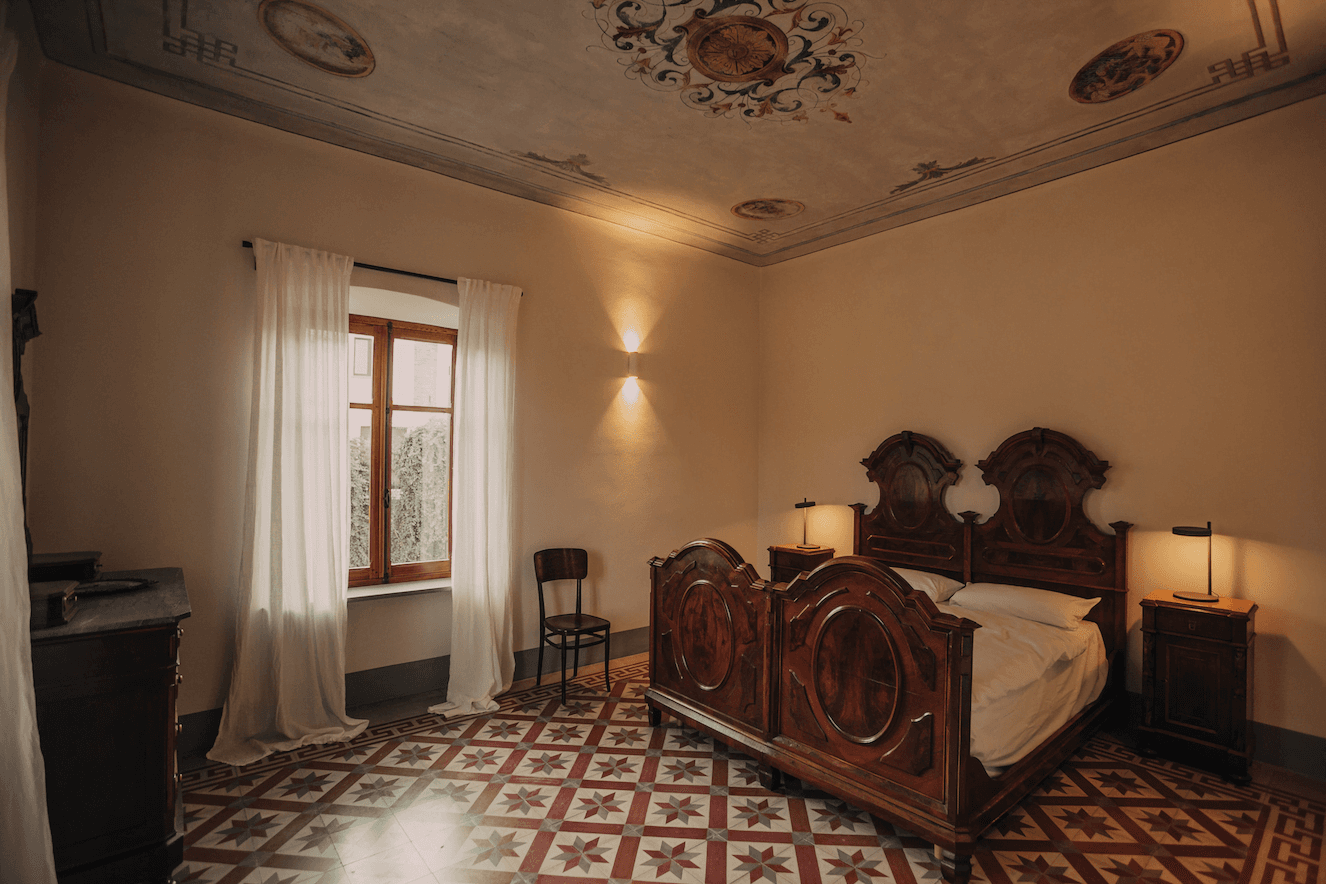 A vintage bedroom features aged tile flooring and an ornate high ceiling. It includes a wooden bed with intricate carvings, two small nightstands with lamps, a wooden chair, and sheer white curtains covering a window that lets in natural light.