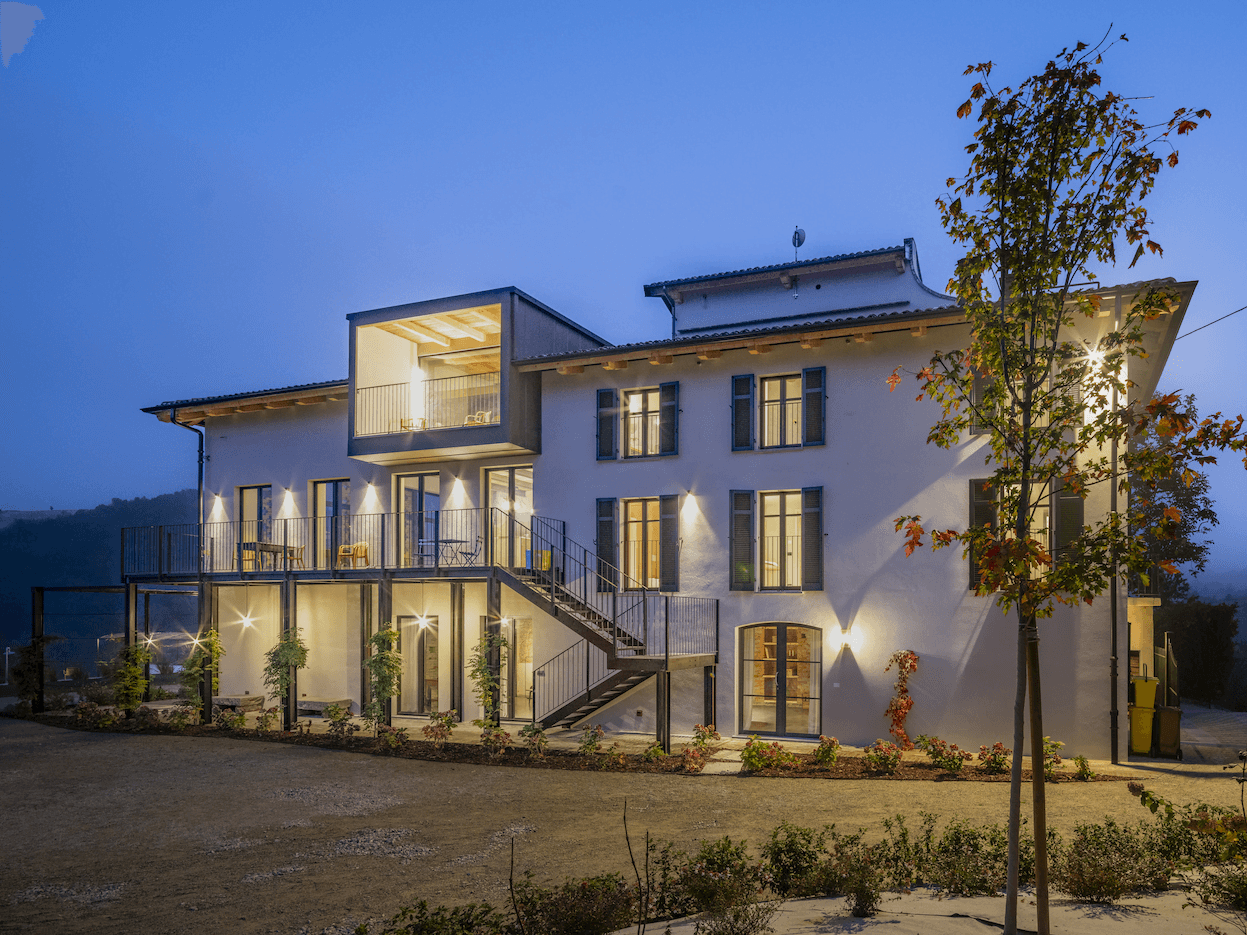 A modern, well-lit two-story house at dusk with illuminated windows. The facade features large glass doors, balconies, and outdoor stairs. The surrounding landscape includes a garden with various plants and a young tree. The sky has a gentle evening blue hue.