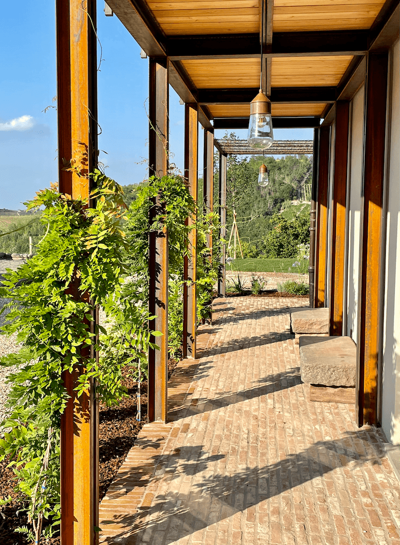 A covered, sunlit patio features a wooden structure with vertical beams supporting a roof. Green vines climb the beams, casting shadows on the brick floor. Large stone seating is arranged along the pathway, offering a view of a lush, green landscape in the background.