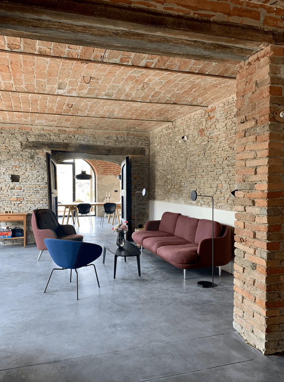 A cozy living space features exposed brick walls and a vaulted ceiling. There is a red sofa, a blue armchair, a floral coffee table, and a modern floor lamp. A wooden table is visible in the background near large open doors. The floor appears to be polished concrete.