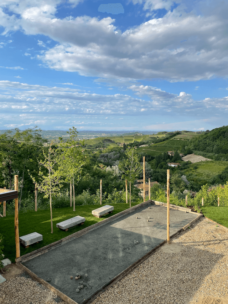A picturesque outdoor space featuring a pétanque court in the foreground with benches and small trees lining the sides. In the background, there are lush green hills, scattered buildings, and a partly cloudy sky.