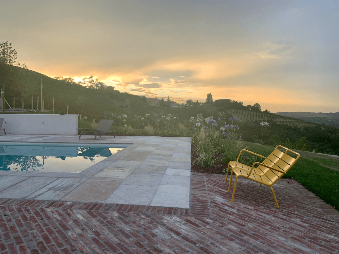 A panoramic view of an area by the pool at sunset features a yellow chair on a brick patio. The pool is surrounded by tiled flooring, with sun loungers and plants nearby. Rolling hills stretch out in the distance beneath a colorful sky.