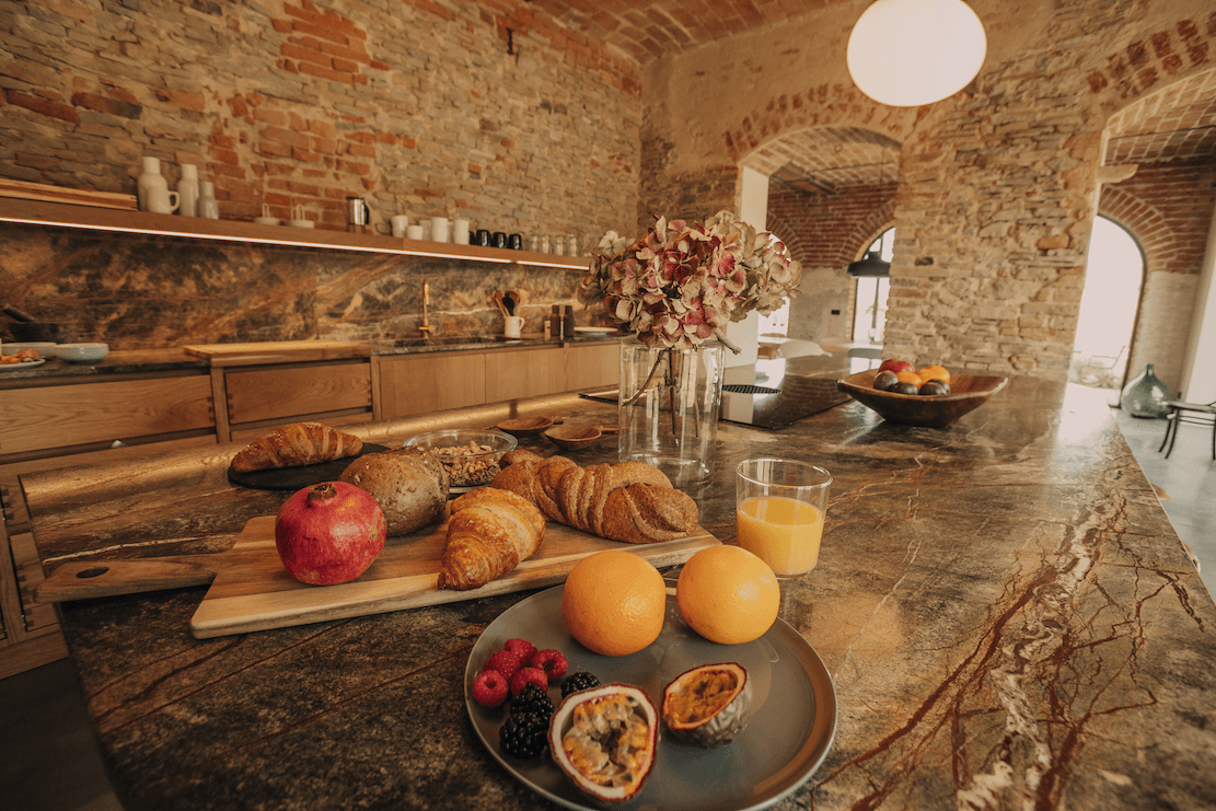A rustic kitchen with a stone wall and wooden cabinets features a marble countertop. On the countertop, there is an assortment of breads on a cutting board, fruits including pomegranates and oranges, a glass of orange juice, berries, and a plate with halved passion fruits.