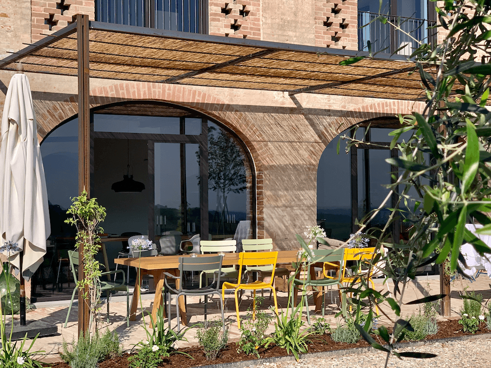 An outdoor patio features a wooden dining table surrounded by chairs in various colors under a pergola. Potted plants and greenery surround the seating area. A glass door with arched frames reflects the landscape and the exterior of the building with brick walls.