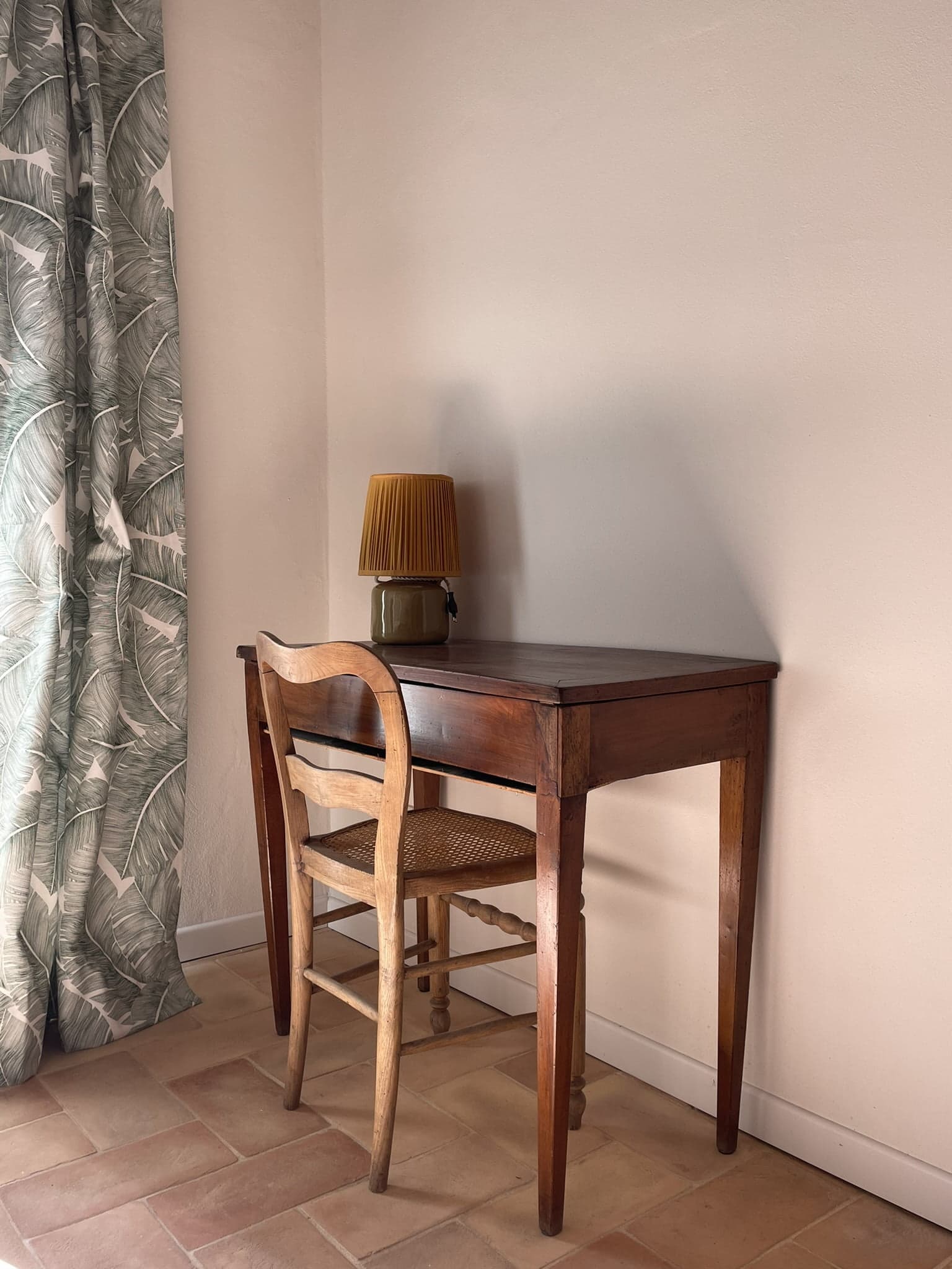 A small wooden table with a wicker chair tucked under it sits against a plain wall. On the table is a lamp with a pleated shade. A curtain with a leaf pattern hangs to the left, and the floor is tiled in a light reddish-brown color.
