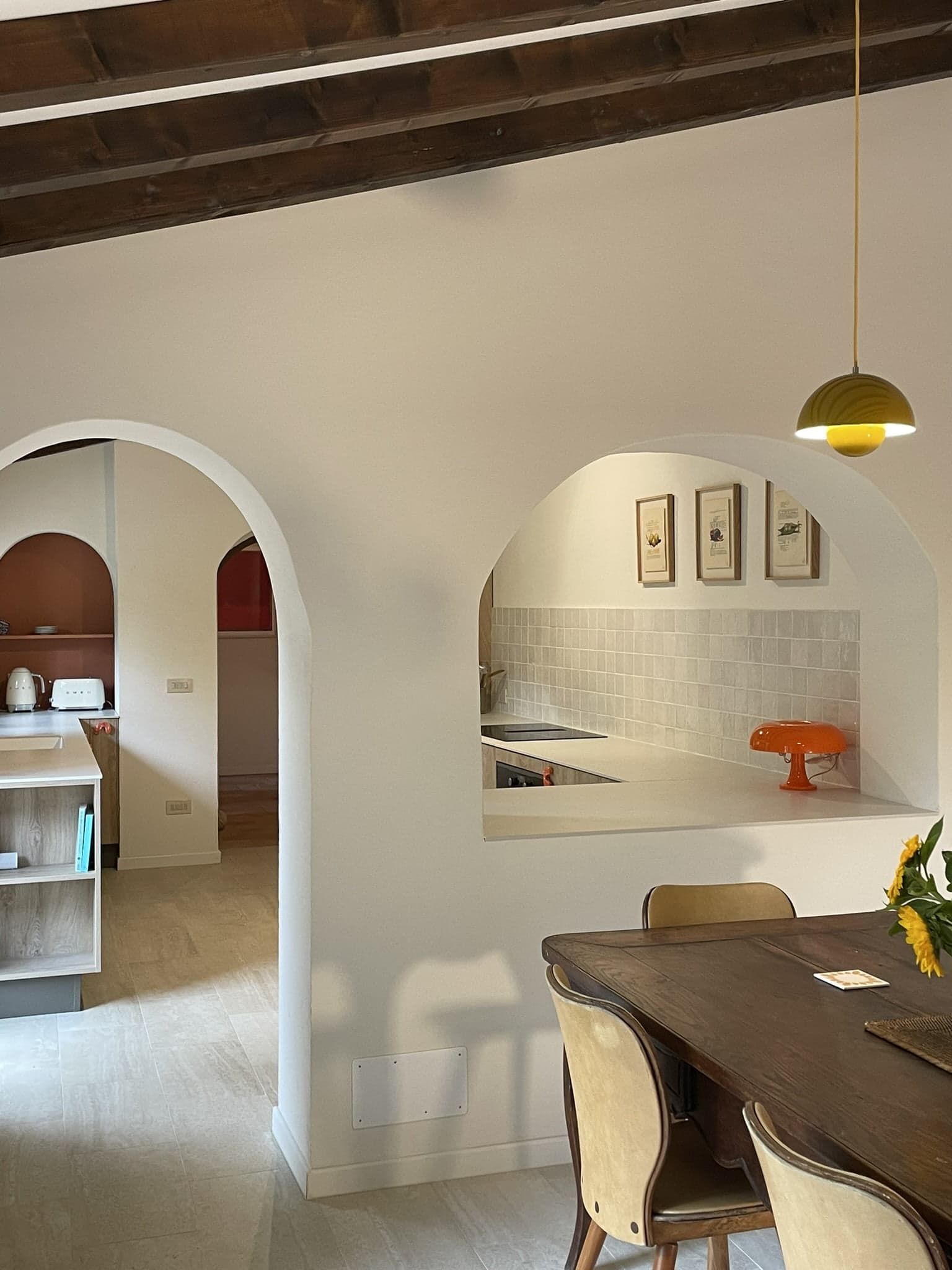 A cozy modern kitchen and dining area with white walls, arched doorways, and a wooden dining table with beige chairs. The kitchen features light tiled backsplash, white countertops, and colorful decor, including a bright orange lamp. Warm lighting adds to the ambiance.