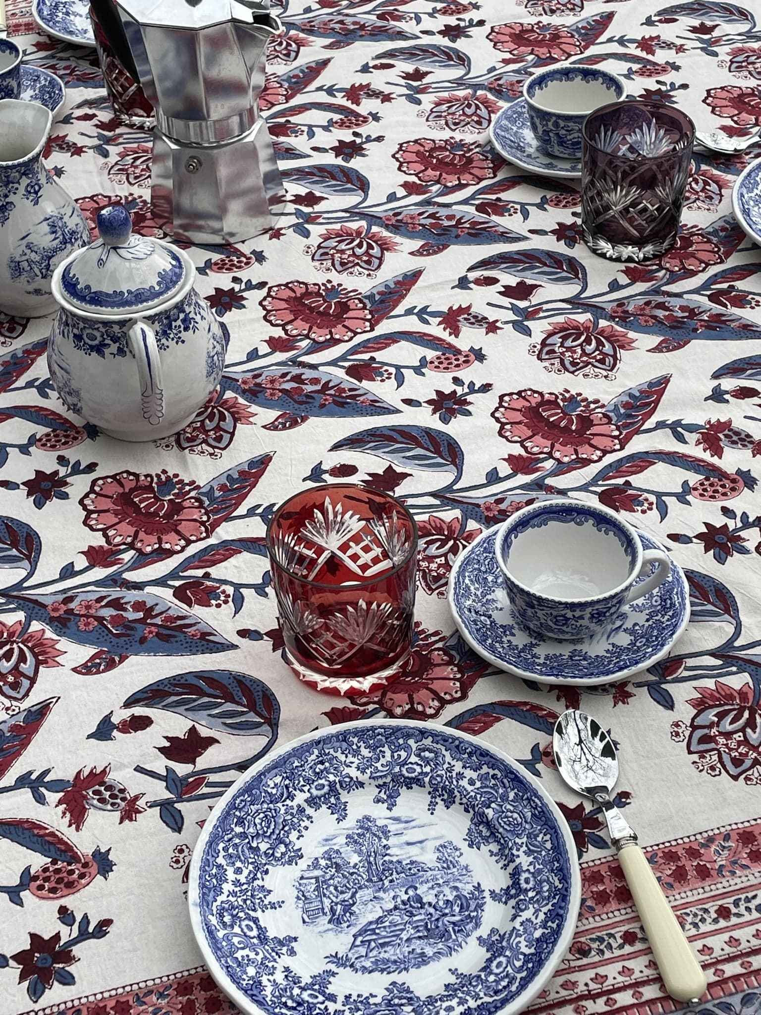 A floral tablecloth holds a tea setting with blue and white ceramic plates, teacups, and a teapot, alongside two colorful crystal glasses and a stovetop coffee maker. A white-handled spoon rests on a saucer beside a teacup.