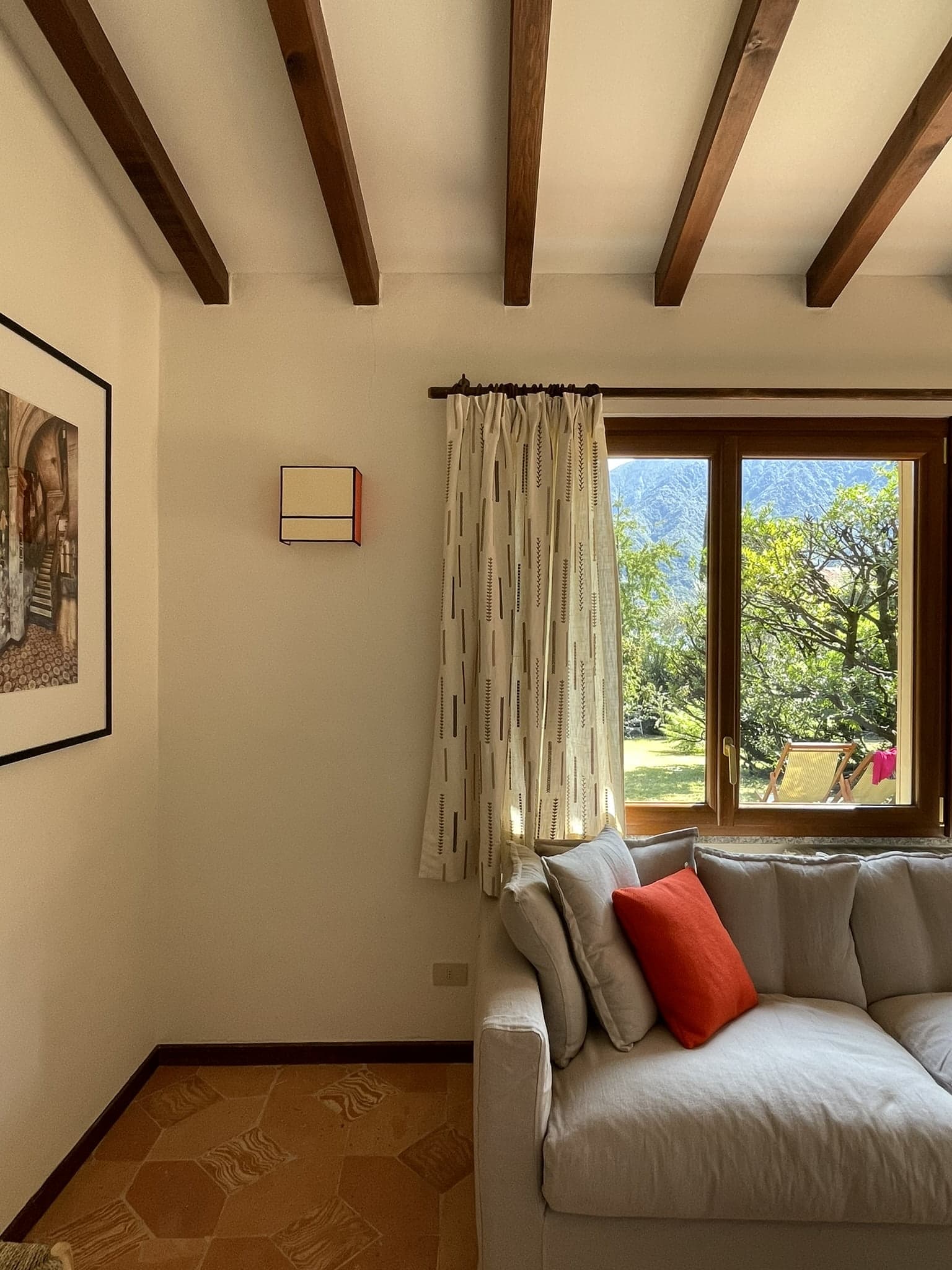 A cozy living room with wooden ceiling beams, a white couch adorned with beige and red cushions, and curtains partially drawn back, revealing a lush garden and mountains through a large window. A framed picture hangs on the white wall to the left.