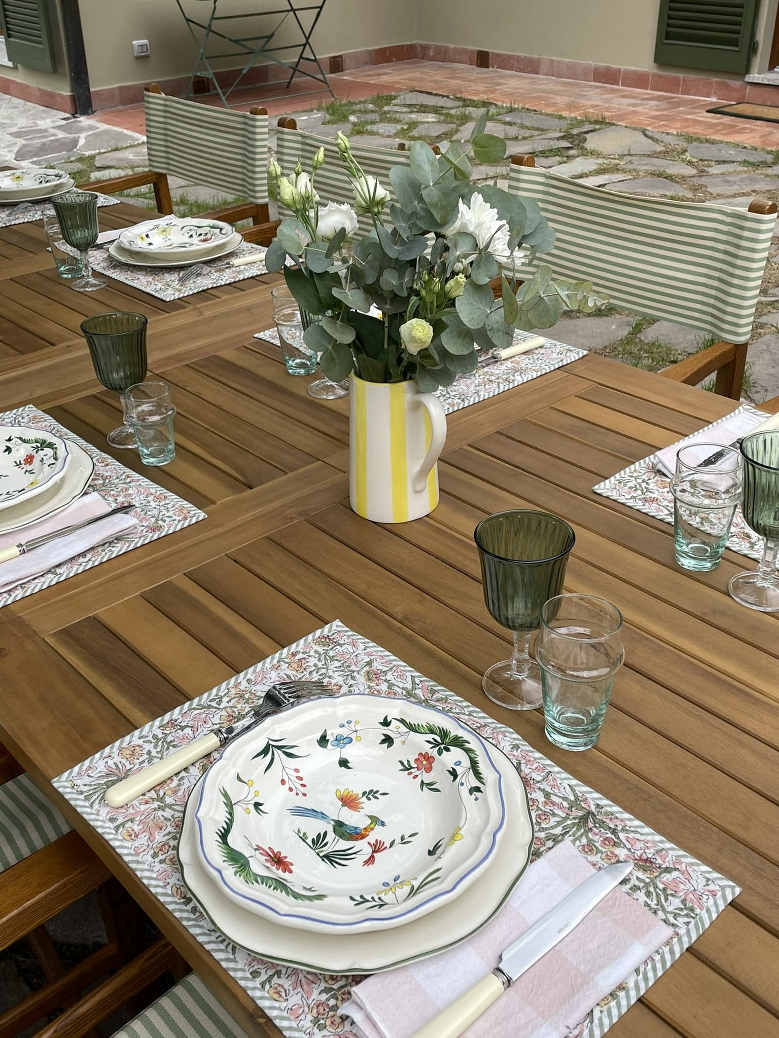 A wooden dining table set outdoors with floral dinner plates, cutlery, green glassware, and napkins. A yellow and white striped vase filled with white and green flowers serves as a centerpiece. The table is surrounded by green and white striped chairs.