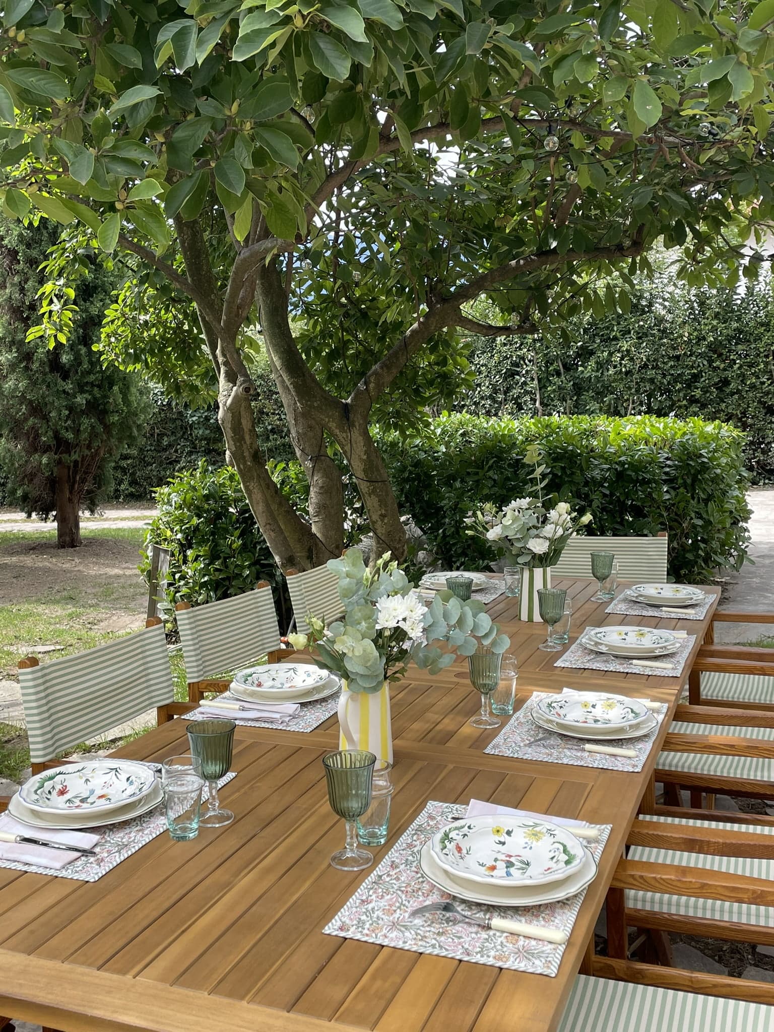 An outdoor dining table set under a leafy tree. The wooden table is adorned with floral plates, glassware, and floral arrangements in vases. Striping on the chairs reflects light green and white. The background features dense greenery and a lawn.