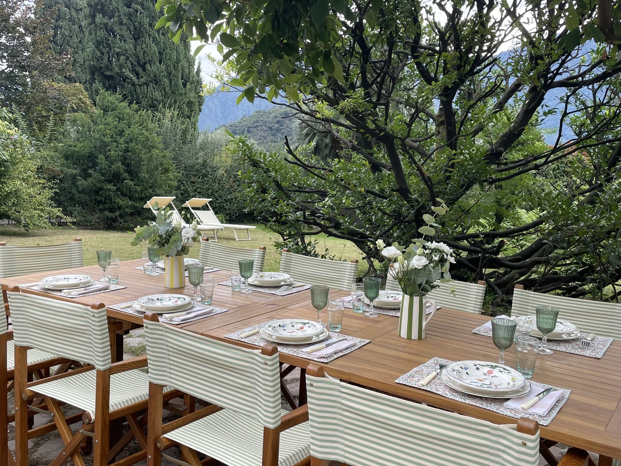 A long outdoor wooden dining table set for a meal with striped chairs, white floral arrangements, and green glassware. The table is situated under a tree in a lush garden with mountains and trees in the background. Two lounge chairs are visible at a distance.
