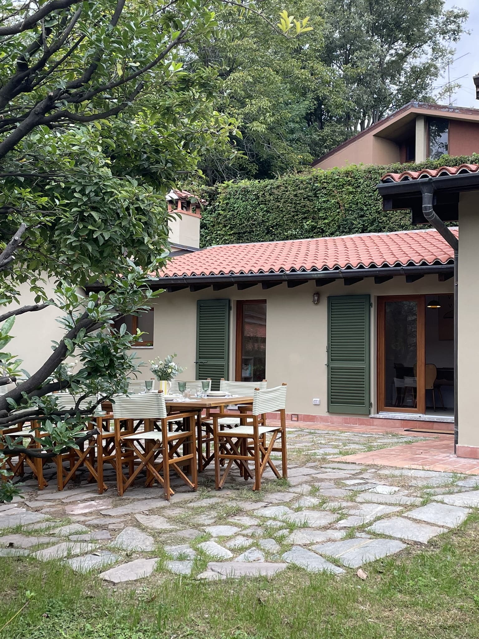 A cozy outdoor patio features a wooden dining table and chairs set on a stone walkway, surrounded by lush greenery. The patio is adjacent to a charming house with green shutters, a red-tiled roof, and large glass doors leading inside.