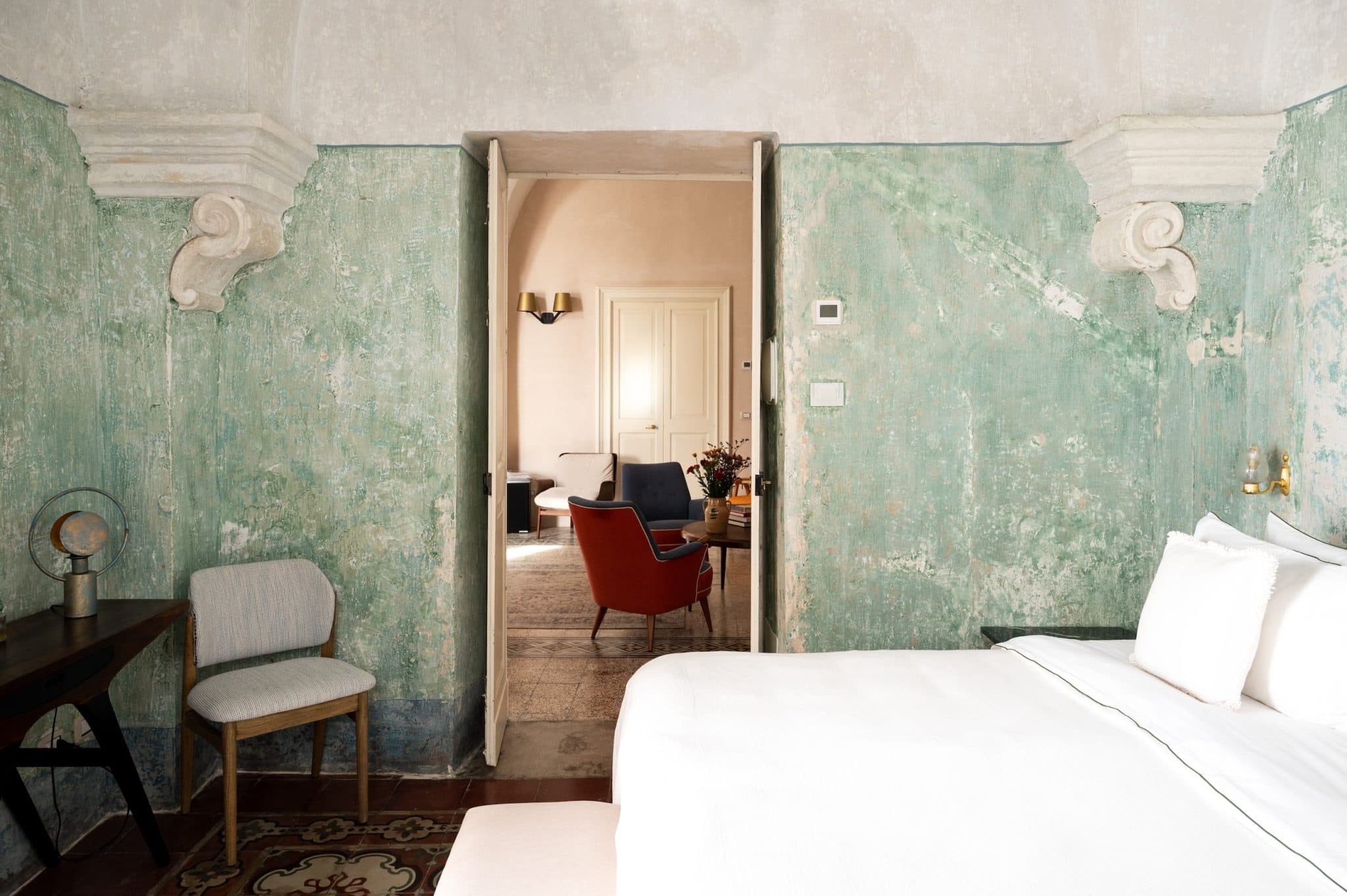 A cozy room with light green, textured walls is seen through an open door. The adjacent room has soft beige walls and features a red armchair, a blue armchair, and a table with flowers. An ornate rug covers the floor. A simple wooden chair is in the foreground.