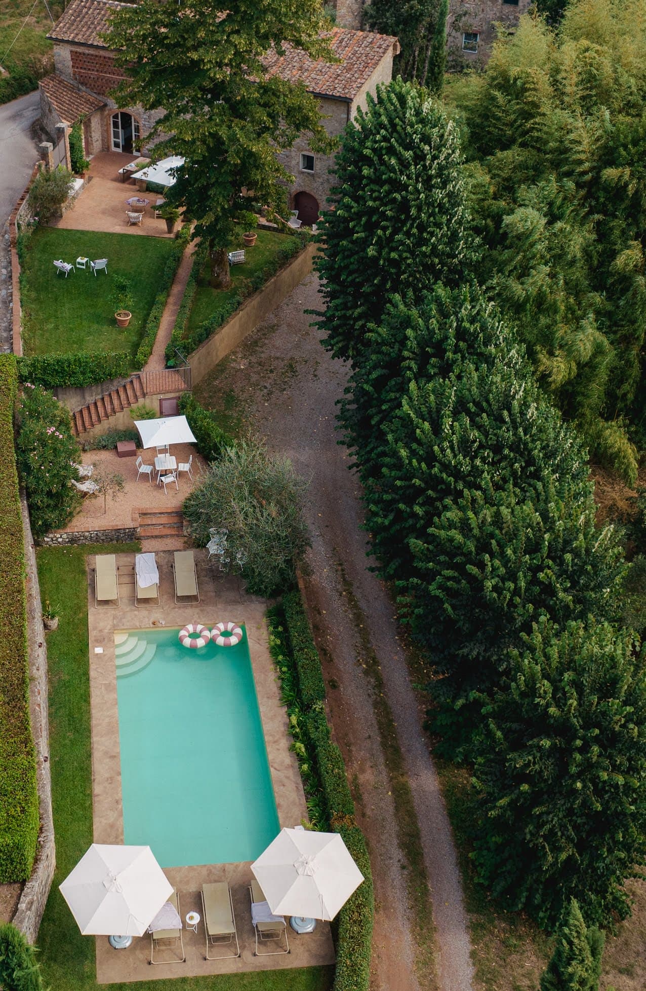 Vue aérienne d'une propriété rustique dotée d'une piscine entourée de chaises longues et de parasols. La propriété comprend une maison en brique avec un toit en tuiles rouges, des arbres verts et des espaces extérieurs pour s'asseoir. Un chemin de gravier longe la piscine.