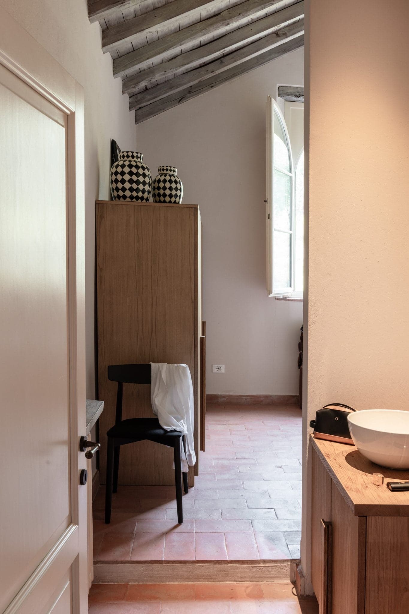 Une pièce au design rustique avec un plafond à poutres apparentes en bois. Une chaise noire recouverte d'un tissu blanc est placée à côté d'un meuble en bois surmonté de vases à motifs noirs et blancs. Une fenêtre au sommet incurvé laisse entrer la lumière naturelle.