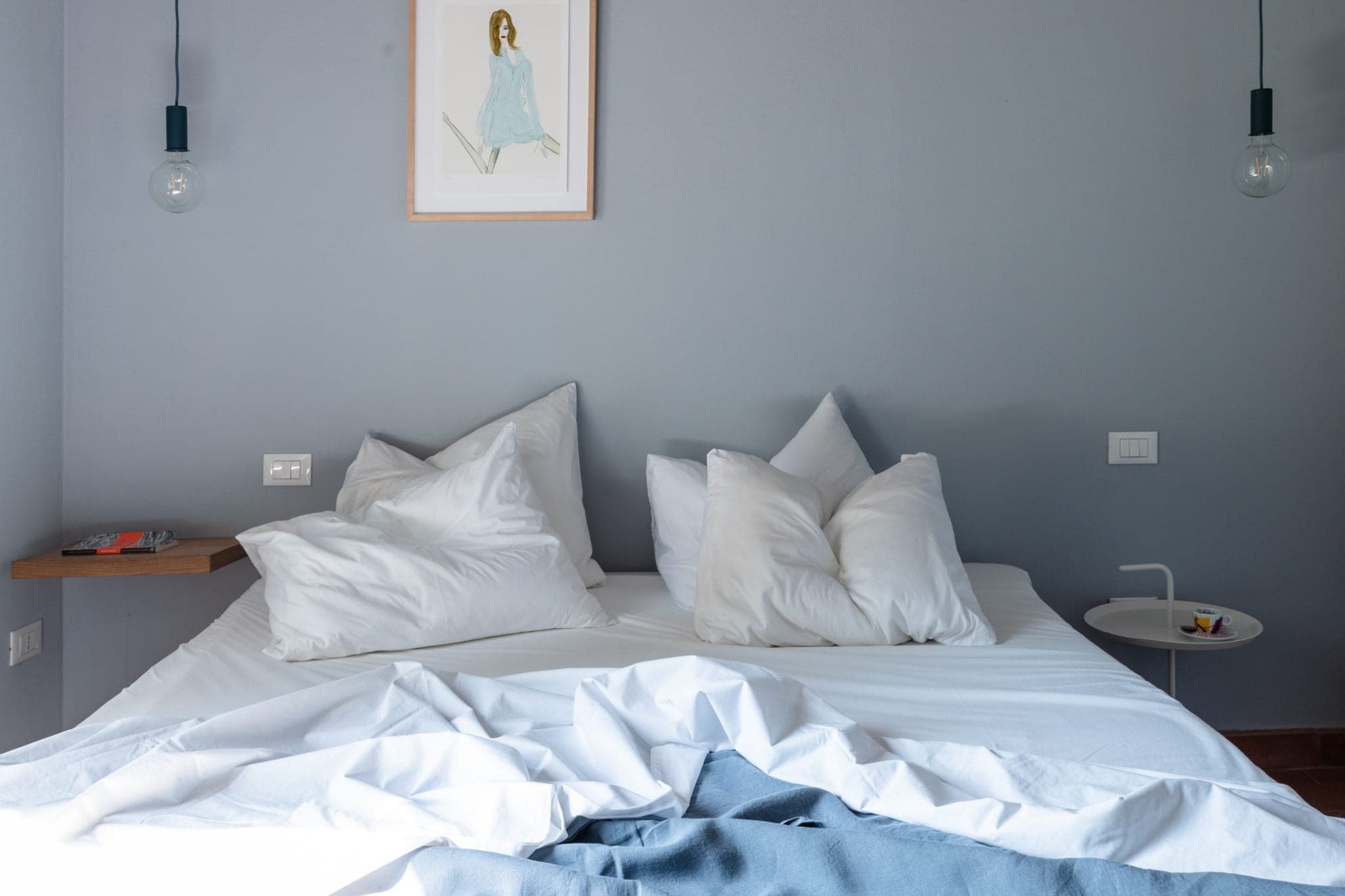 Une chambre moderne avec un lit double soigneusement fait avec des oreillers et des draps blancs, une petite table d'appoint ronde blanche avec une lampe et quelques objets, et un dessin d'une personne en tenue bleue accroché au mur gris clair au-dessus du lit.